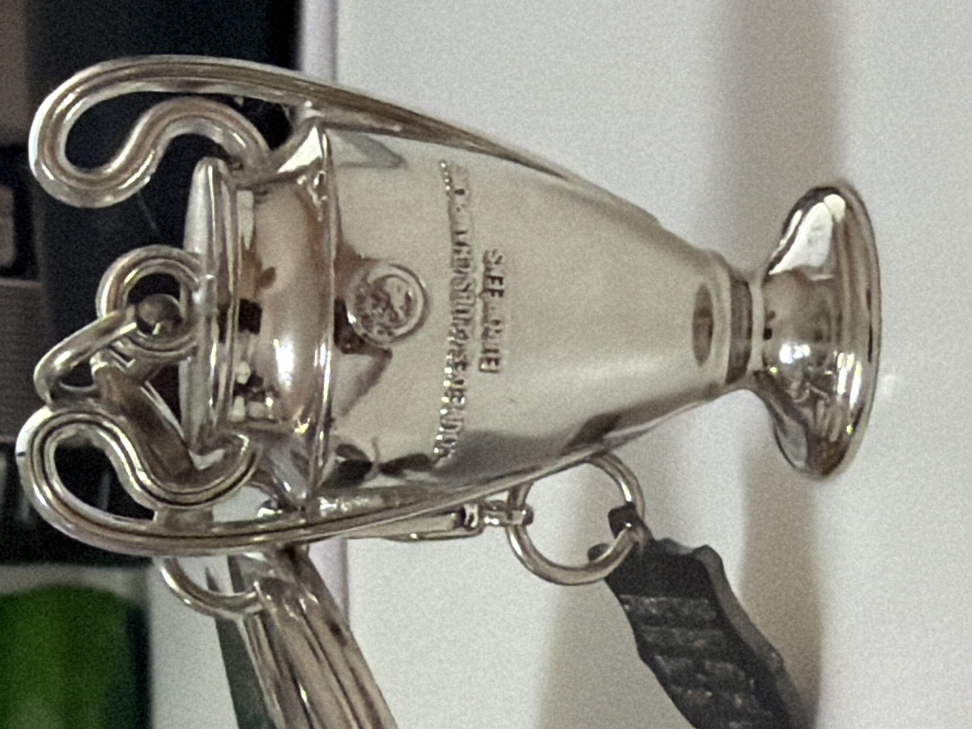The image shows a close-up of a silver trophy keychain, likely representing the UEFA Champions League trophy, placed on a white surface. The trophy is detailed with engraved text that reads "WINNERS OF THE EUROPEAN CUP" and "EUROPEANS." There are also circular emblems on the trophy. A dark, irregular shaped object, possibly a piece of a broken keychain or charm, is attached to the trophy by a metal ring. Another metal ring connects the trophy to a set of keys, one of which is partially visible. The background is blurred, with a hint of a green object on the left and dark areas on the right, suggesting an indoor setting. There are no people present in the image. The location in the city of Kyiv, Ukraine, is not discernible from the image content.