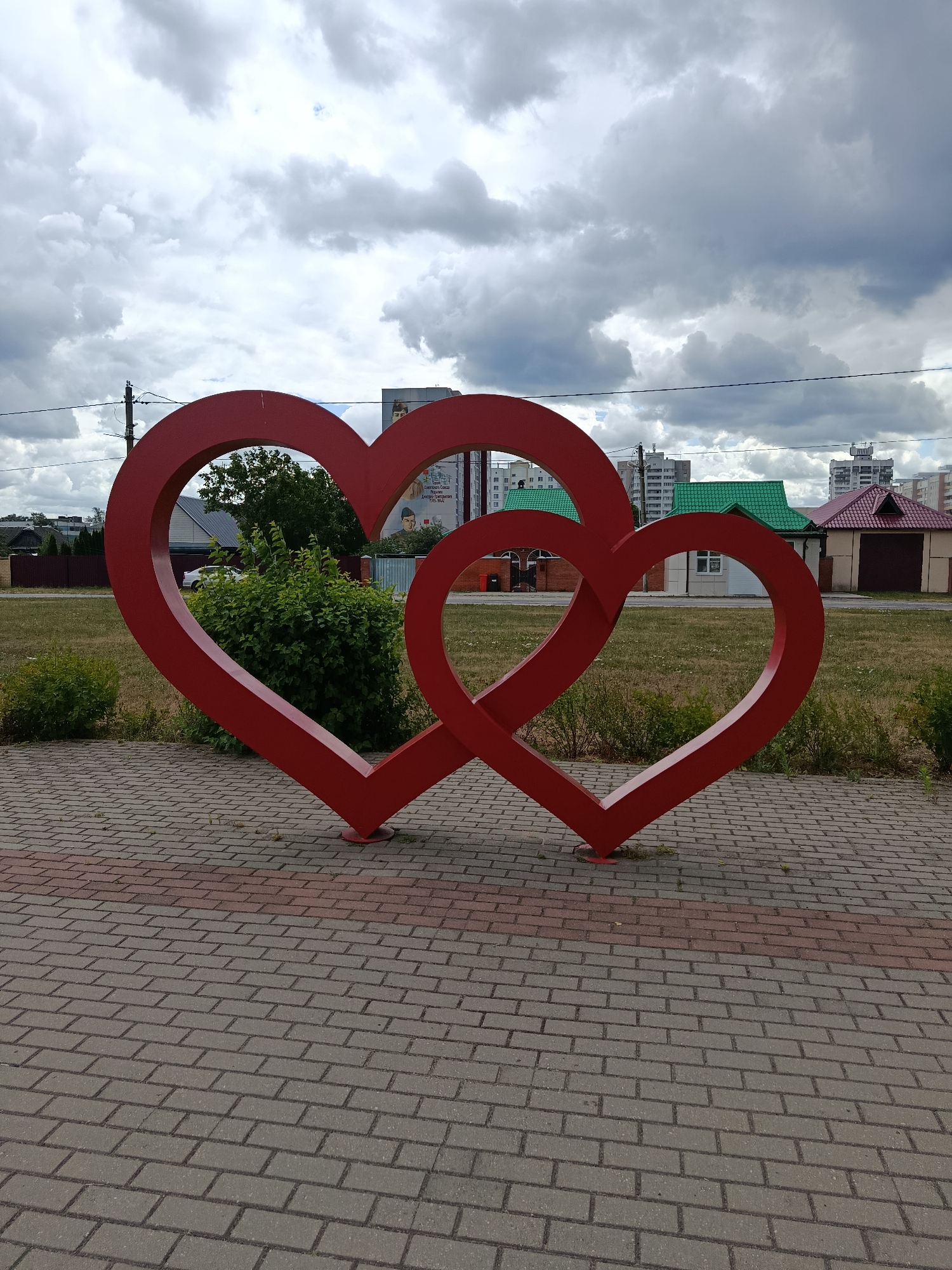 A close-up, low-angle shot captures two large, red heart sculptures intertwined in the foreground. They are positioned on a paved walkway made of grey and red rectangular bricks. Behind the sculptures, a grassy area is dotted with green bushes. In the background, several buildings are visible, including houses with green and burgundy roofs and taller, multi-story buildings further in the distance. The sky above is filled with dramatic, grey, and white clouds, suggesting an overcast or partly cloudy day. There is no clear indication of the time of day, but the lighting suggests it is daytime. No people are visible in the image. A billboard with an image of a man and some text in Cyrillic script is partially visible on a building in the background. The scene appears to be an outdoor public space, possibly a park or a town square, in Svietlahorsk, Belarus. The overall mood is somewhat somber due to the cloudy sky, but the prominent heart sculptures add a touch of romance or symbolism to the scene.