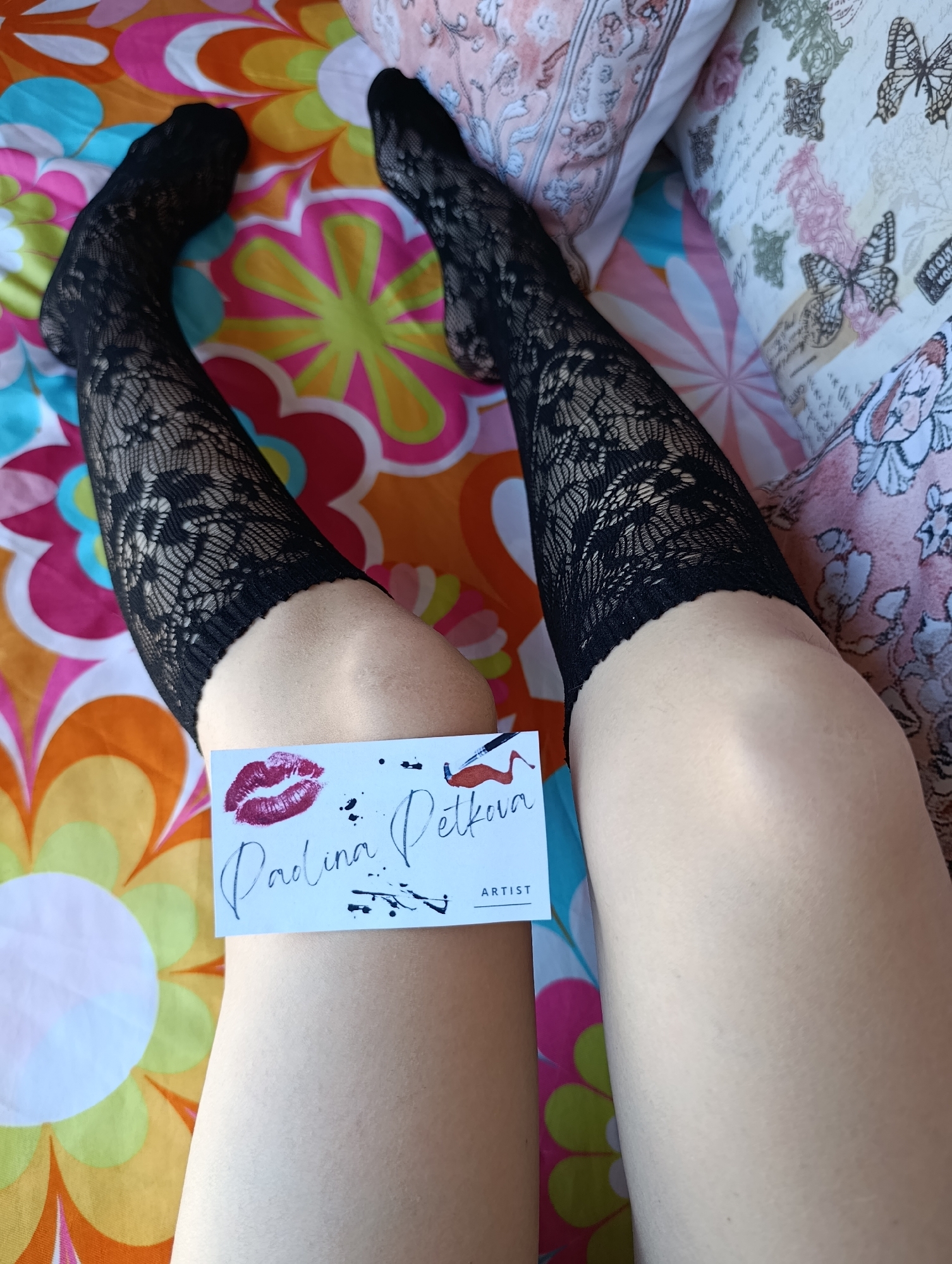 The image presents a view of a person's legs, clad in black lace stockings. The person is sitting on a bed with a colorful floral patterned bedspread and pillows. One pillow has a butterfly pattern. A white card is held on the left leg, displaying the name "Paulina Petkova" and the word "Artist," along with a lipstick kiss and a drawing of an applicator with smeared product. The setting appears to be indoors, potentially a home. There is no information to determine if this photo was taken in Svilengrad, Bulgaria. The image lacks context to definitively link it to Svilengrad.