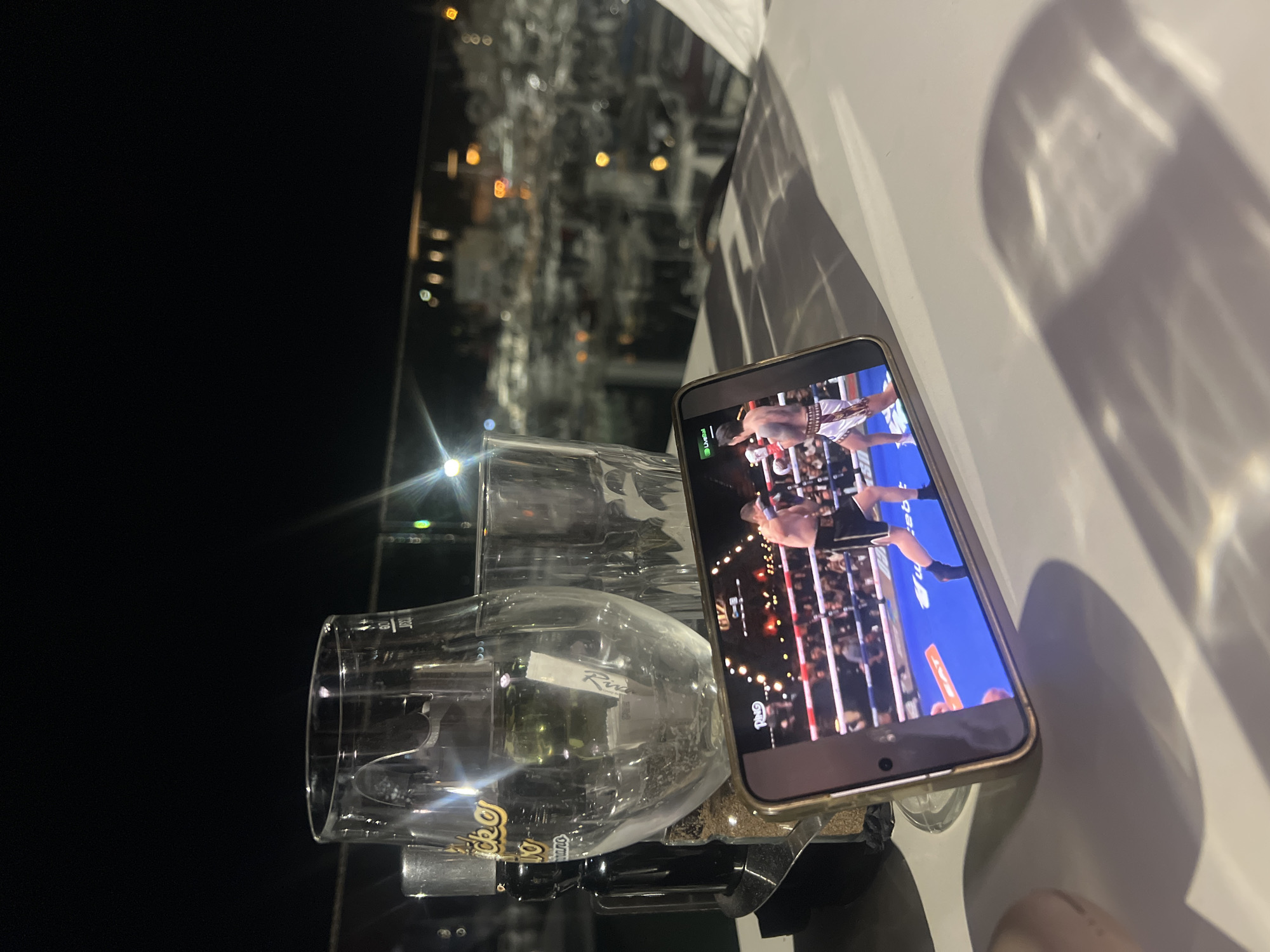 The image depicts a smartphone resting on a surface, displaying a boxing match. Next to the phone are two stacked clear glasses. The background reveals an out-of-focus night scene with numerous lights, suggestive of an urban environment, consistent with being in the city of Budva, Montenegro. The light source from the distance suggests a coastal location. The surface the phone and glasses rest on is light-colored. A partial view of a person's finger appears in the bottom right corner of the image.