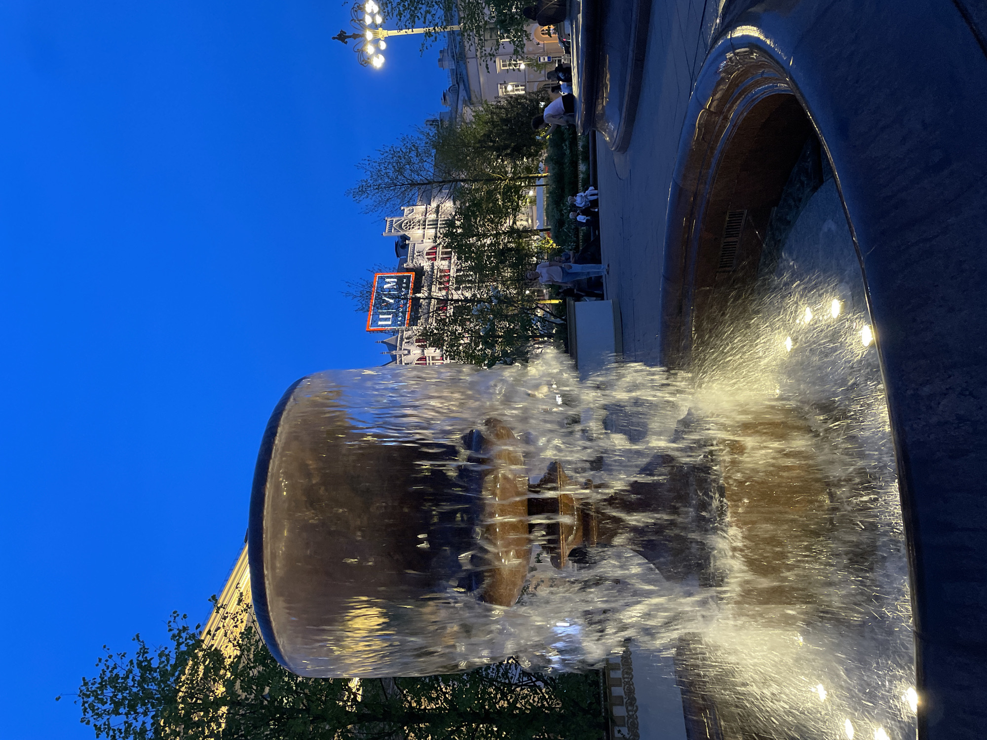 The image depicts the Cascade Fountain in Gorky Park, Moscow, Russia, at dusk. The fountain features a multi-tiered bowl design, with water cascading down the sides. The water is illuminated by lights, creating a sparkling effect. In the background, buildings can be seen, including one with a prominent "UM" sign in Cyrillic script and English letters, suggesting it's a department store called "TSUM" or similar. The sky is a deep blue, indicating twilight. People are visible in the background, appearing as indistinct figures enjoying the park. Trees are present in the park, adding greenery to the scene. The photograph is taken from a low angle, emphasizing the fountain and capturing its details.
