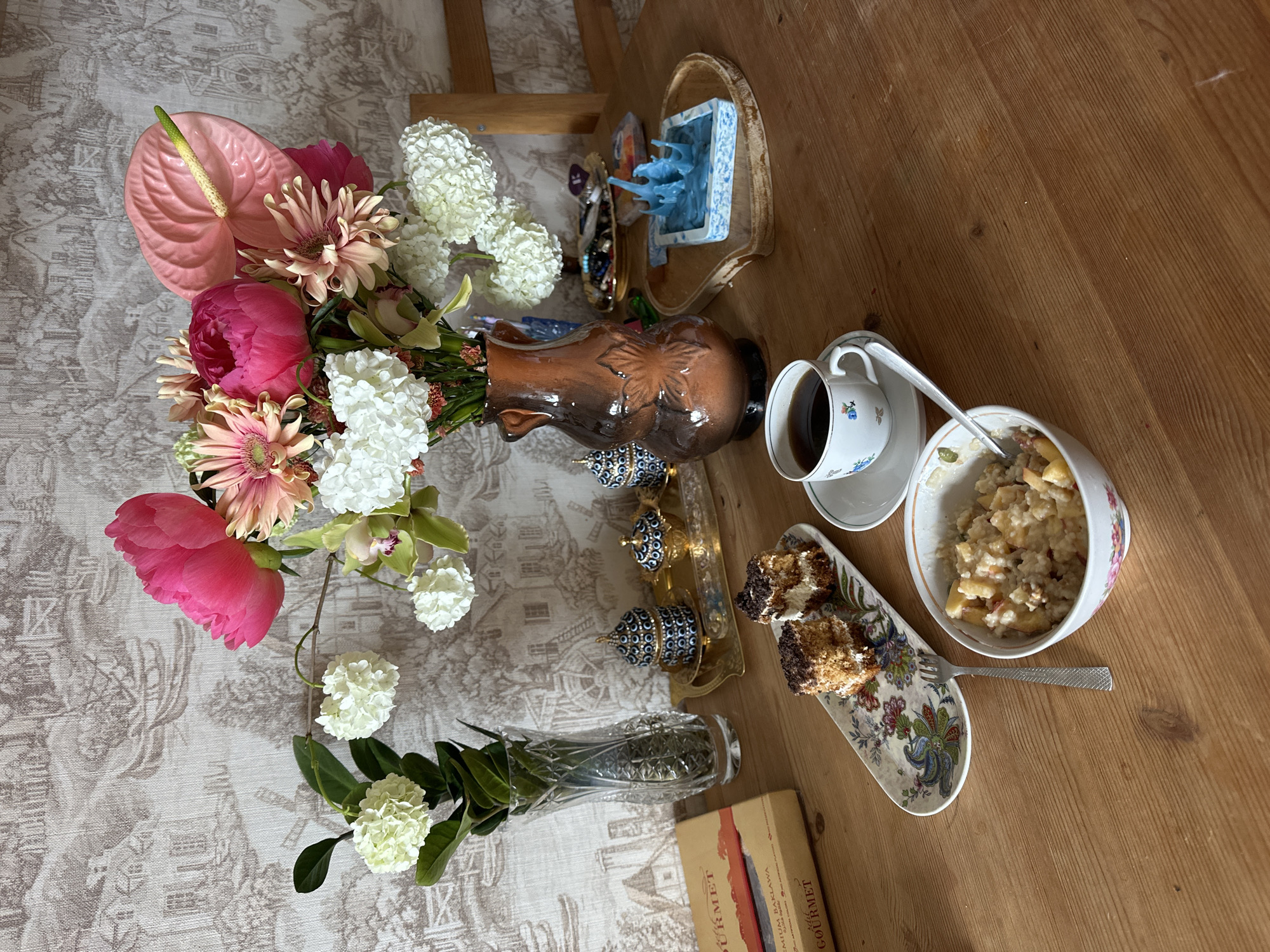 The image shows a still life scene on a wooden table. The table is partially covered with a patterned tablecloth. On the table are several items including:
- A vase filled with a variety of flowers including pink peonies, anthuriums, gerberas, and white hydrangeas
- A brown ornamental vase
- Two small ornate containers on a golden tray
- A cup of coffee on a saucer
- A decorative plate holding pieces of cake
- A bowl of porridge or oatmeal with a fork
- A crystal vase holding a green leafy plant
- A box labeled "Gourmet"
The scene appears to be indoors, possibly in a home in Vladimir, Russia. The context is inferred from the box's mention of "Baklava", a desert typical of Russian cuisine. No people are visible in the image.