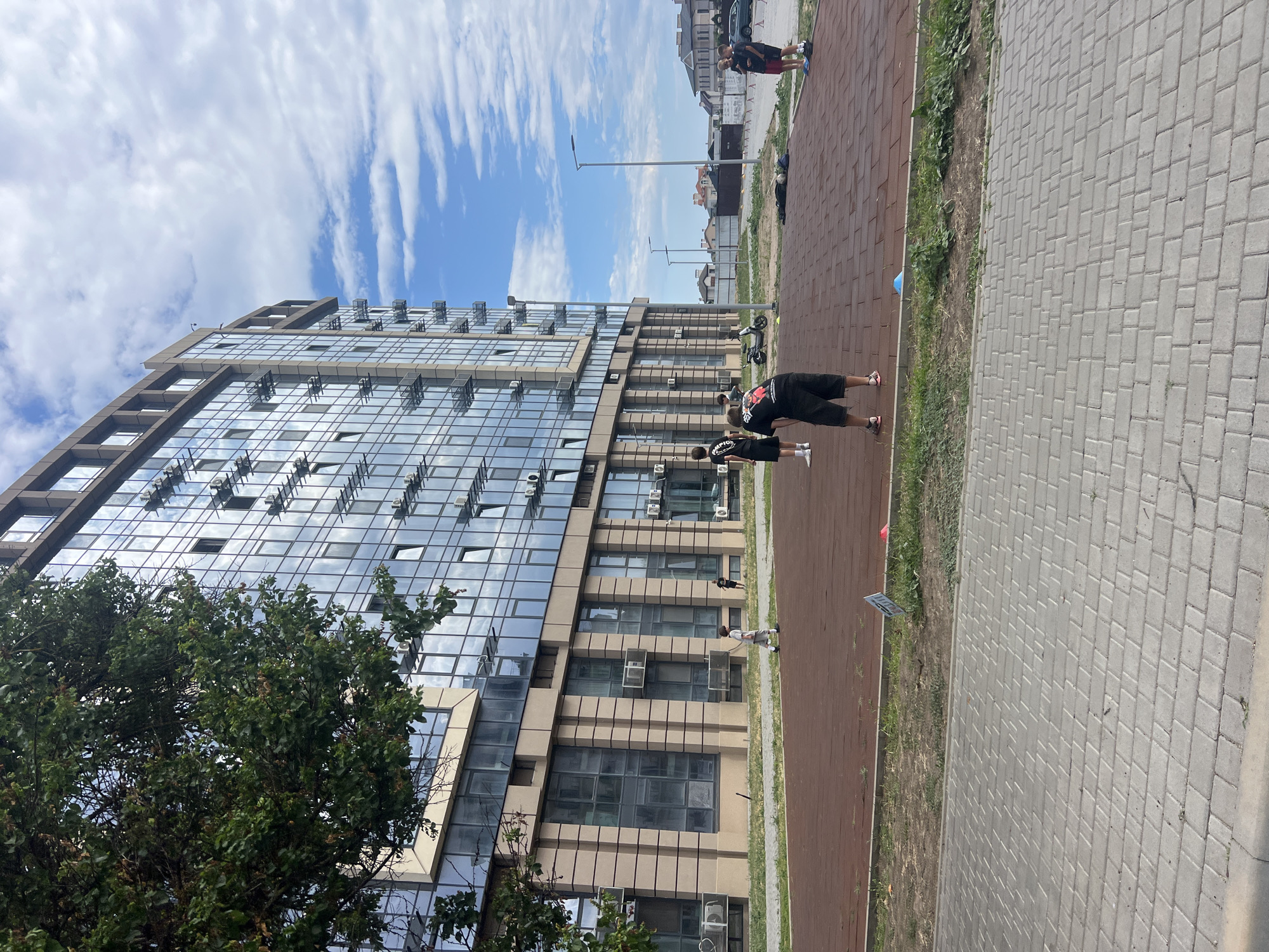 In Fontanka, Ukraine, a group of individuals are gathered on a red-surfaced sports court in front of a modern, glass-facade building. The building features numerous windows and external air conditioning units. Several people are standing and some appear to be playing a game, with one person in the foreground wearing a black t-shirt and shorts, and another in a white t-shirt and shorts. There is a scooter parked near the building. The sports court is bordered by a grassy area and a paved walkway. The sky above is partly cloudy with a bright blue expanse. In the background, residential buildings and a fence can be seen.