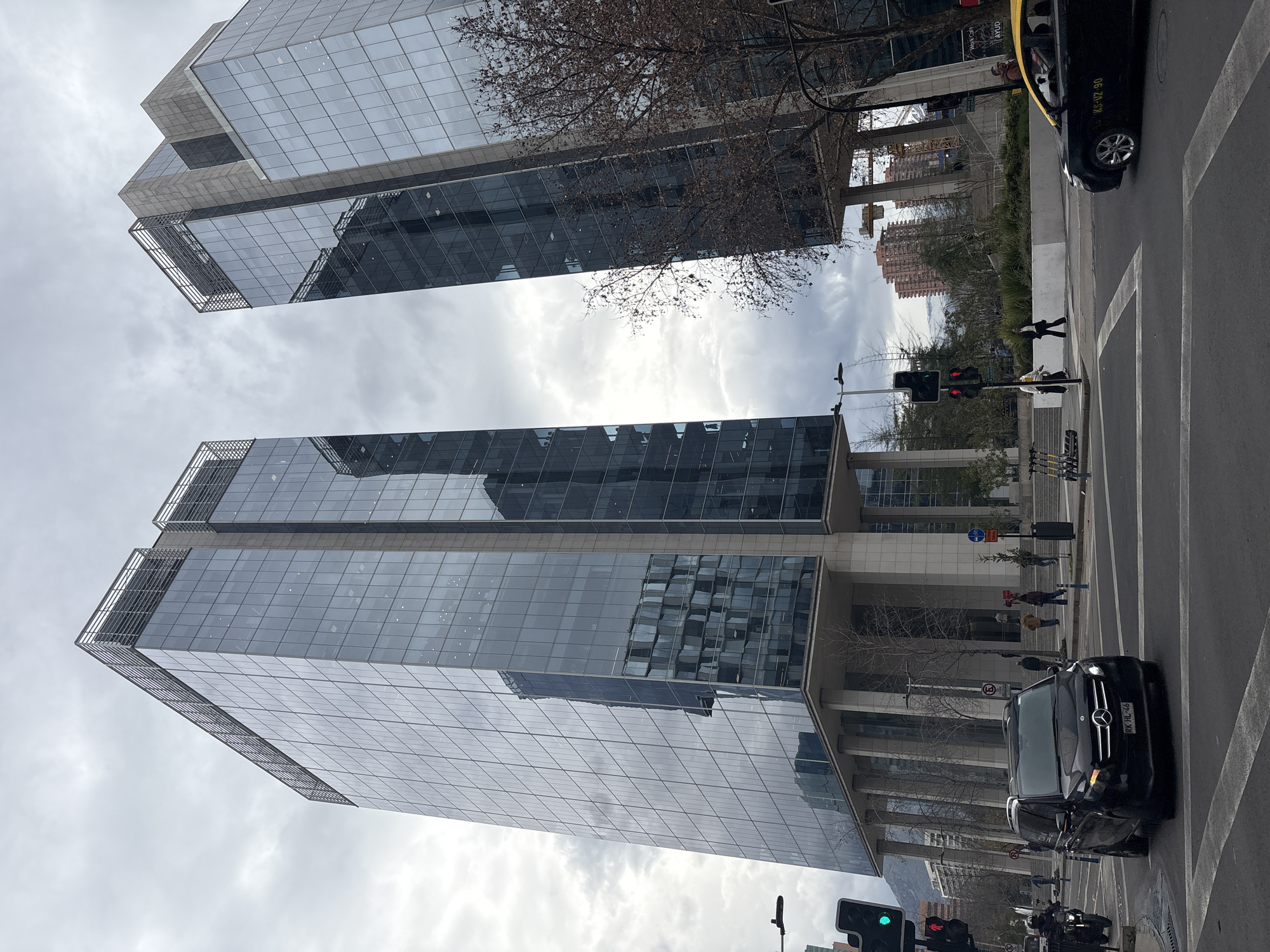 The image captures a street-level view of modern skyscrapers in Santiago, Chile, under an overcast sky. The foreground shows a street intersection with traffic lights, lane markings, and a black Mercedes-Benz Vito van turning left. A yellow taxi is visible in the right lane. Pedestrians can be seen on the sidewalks, and a motorcycle is present to the left. The two prominent glass-fronted office buildings dominate the background. The building on the left is taller and features a reflective facade displaying geometric patterns of reflected buildings. The building on the right is slightly shorter and also has a glass exterior. Bare trees with brown foliage are interspersed between the buildings and the street. In the distance, more buildings and a crane are visible, suggesting ongoing construction or development in the area. The overall atmosphere is urban and functional.