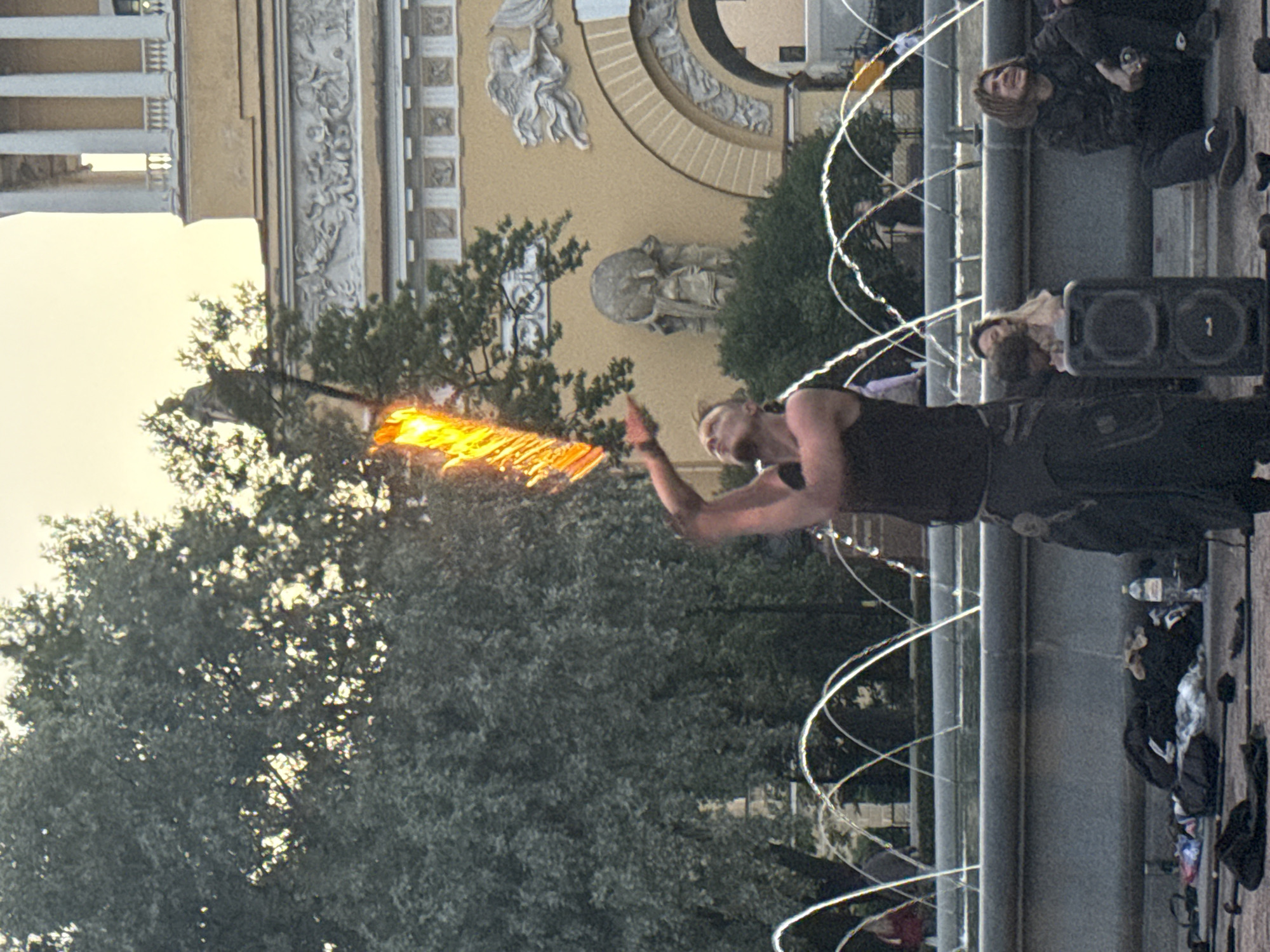 A male fire performer is shown in the center of the image, tossing a flaming object that appears to be a baton or staff. He is wearing a dark sleeveless shirt and dark pants with a patterned belt. In front of him is a large fountain with water arching from multiple spouts. Behind the fountain is a lush green tree and a building with classical architecture, including columns and decorative carvings. To the right of the performer, two people are sitting on the edge of the fountain. One is a young man with long, light-brown hair, wearing a dark jacket and sitting with his legs extended. The other person is partially obscured, but appears to be a young woman. In the foreground, several bags, clothing items, and what appear to be performance props are scattered on the ground. The sky in the background is a pale orange, suggesting it is either sunrise or sunset. This scene is likely taking place in a public square in Saint Petersburg, Russia, possibly near the Admiralty building or Palace Square, given the architectural style.