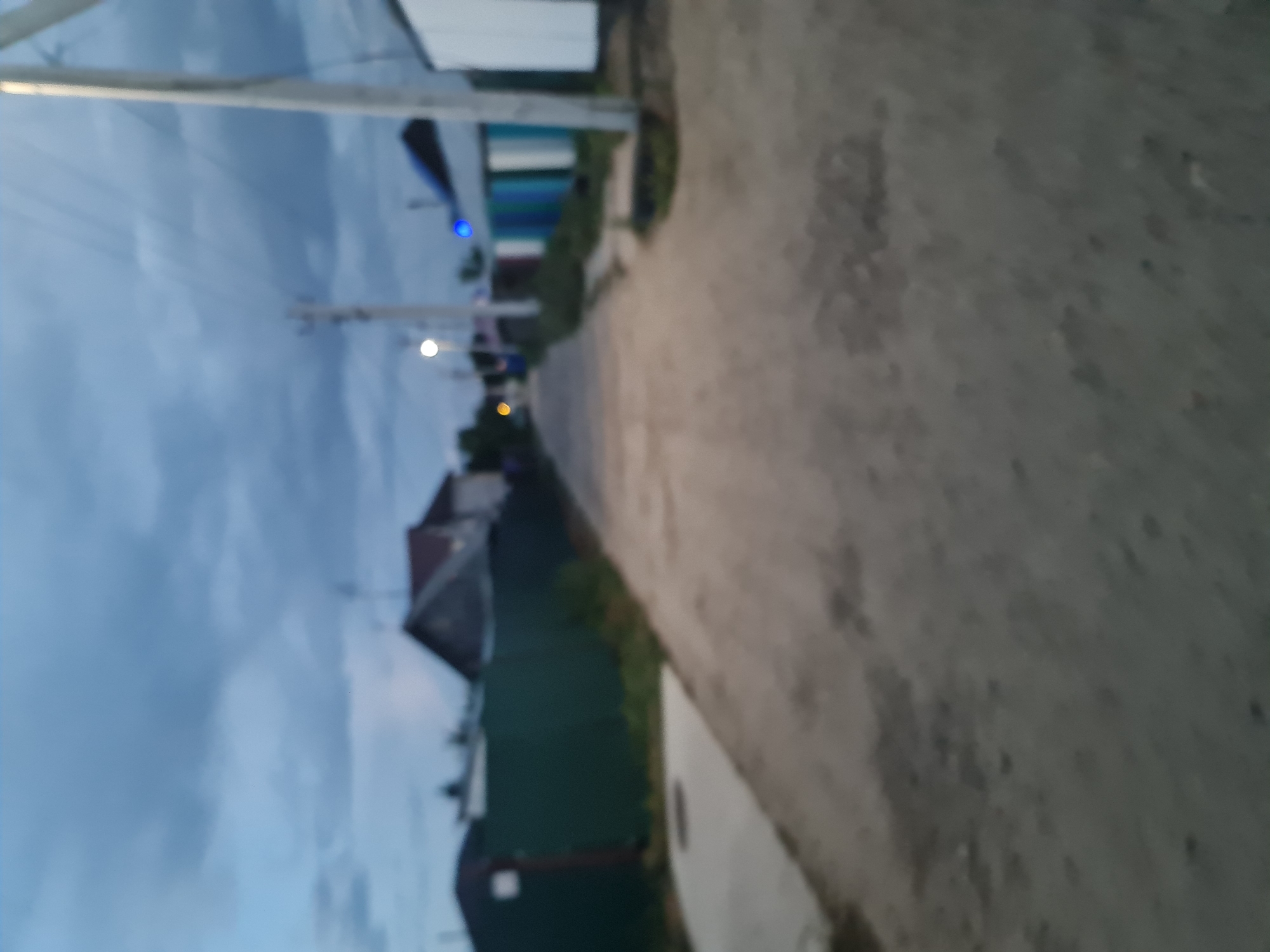 The image depicts a blurred view of a street in a rural or suburban area of Angarsk, Russia. The scene appears to be at dusk or twilight, with a cloudy sky overhead. On the left side of the street, a dark green fence runs along the property line of houses. A paved sidewalk is visible next to the fence. The street itself is unpaved, appearing to be dirt or gravel, with some unevenness.
In the distance, houses with angled roofs are visible, one of which has a prominent dark roof. There are also several outbuildings or garages with multi-colored siding, including shades of blue and green. Utility poles with power lines are present along the street, and a street light is illuminated. A faint blue light, possibly from a house or vehicle, is also visible. No people are present in the image. The overall impression is one of a quiet, residential street in a provincial Russian town.