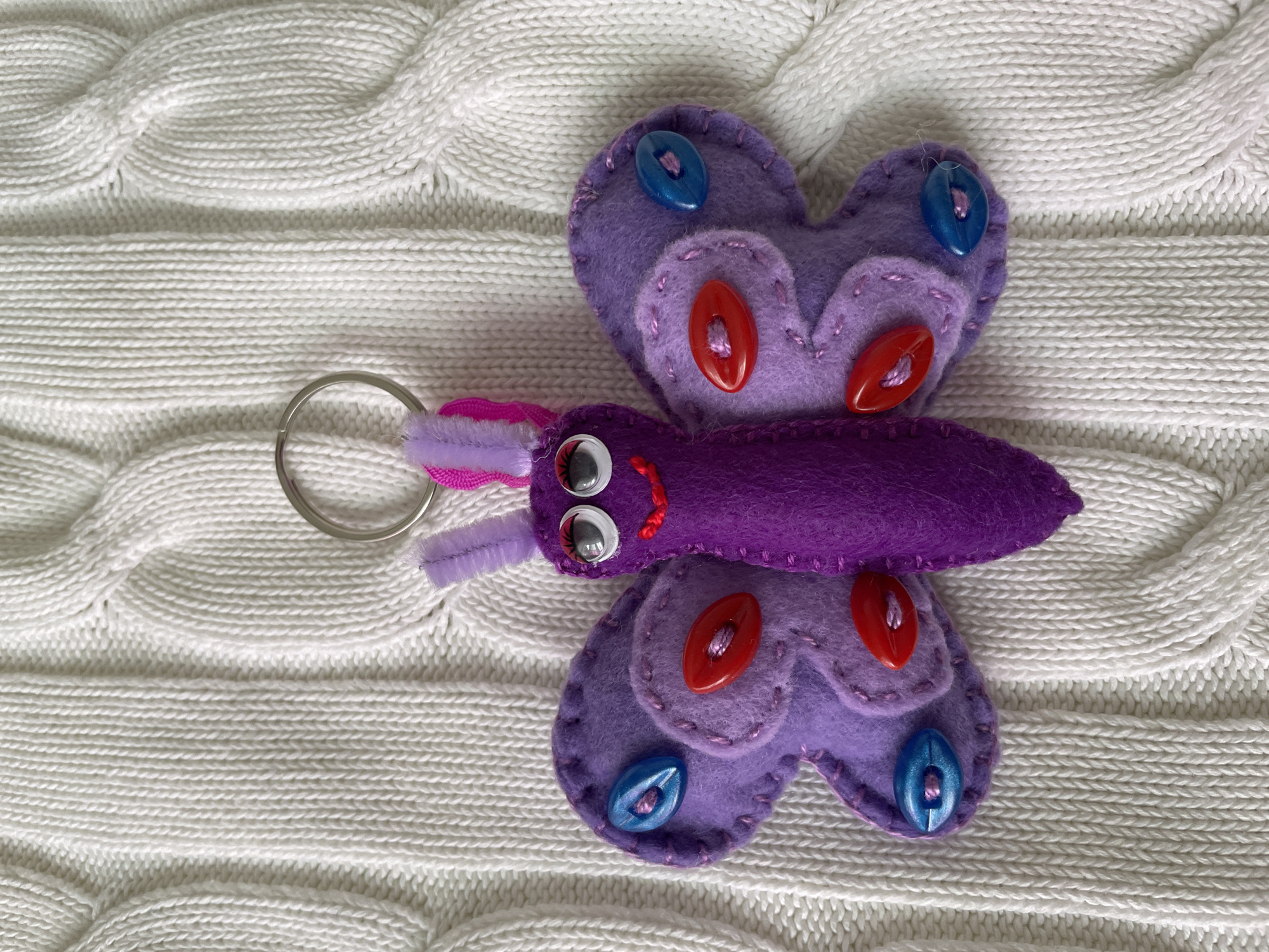 A top-down shot shows a handmade purple butterfly keychain on a white knitted background. The butterfly's wings are heart-shaped and adorned with blue and red buttons. The body is a darker shade of purple, with googly eyes and a small red smile. A purple and pink fuzzy pipe cleaner forms the antennae. A silver keyring is attached to the butterfly. The knitted background has a cable-knit pattern.