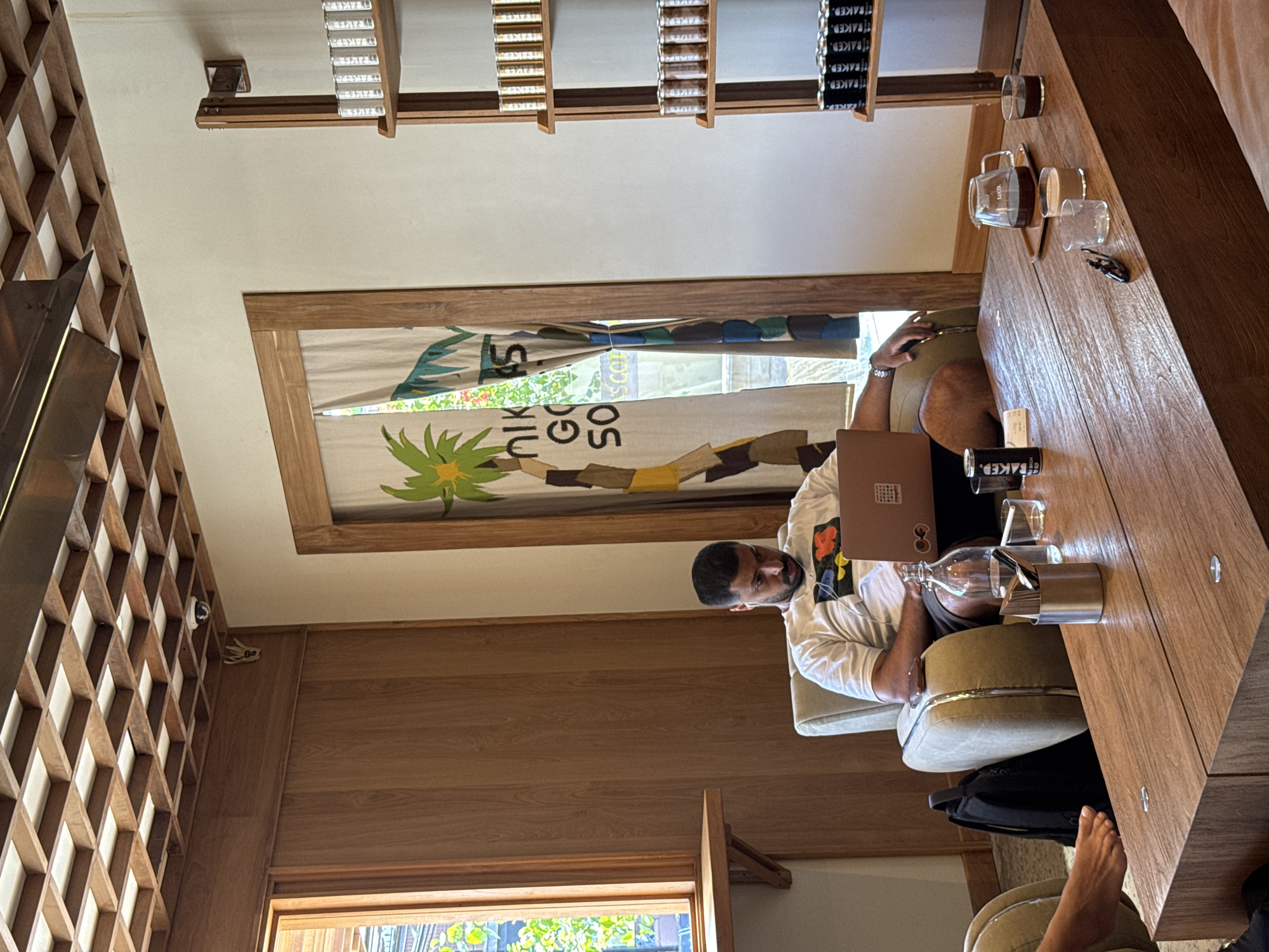 Here is a detailed description of the image:
The image appears to be taken inside a cafe in North Kuta, Indonesia. A man is sitting at a wooden table using a laptop. On the table, there is a coffee cup labeled "FAKED", along with glasses, a water bottle, and a metal container holding cutlery. The man is wearing a white t-shirt with a colorful design and has a beard. He seems to be looking at the laptop screen. Below the table, there's a black bag and part of a bare foot visible.
Behind the man, there's a window with a decorative frame featuring a palm tree design and the words "NIK GO SO" partially visible. Above the window, there are shelves with various products labeled "FAKED". The cafe interior consists of wooden walls and a lattice-like design element on the left side. The lighting suggests a bright, daytime environment.