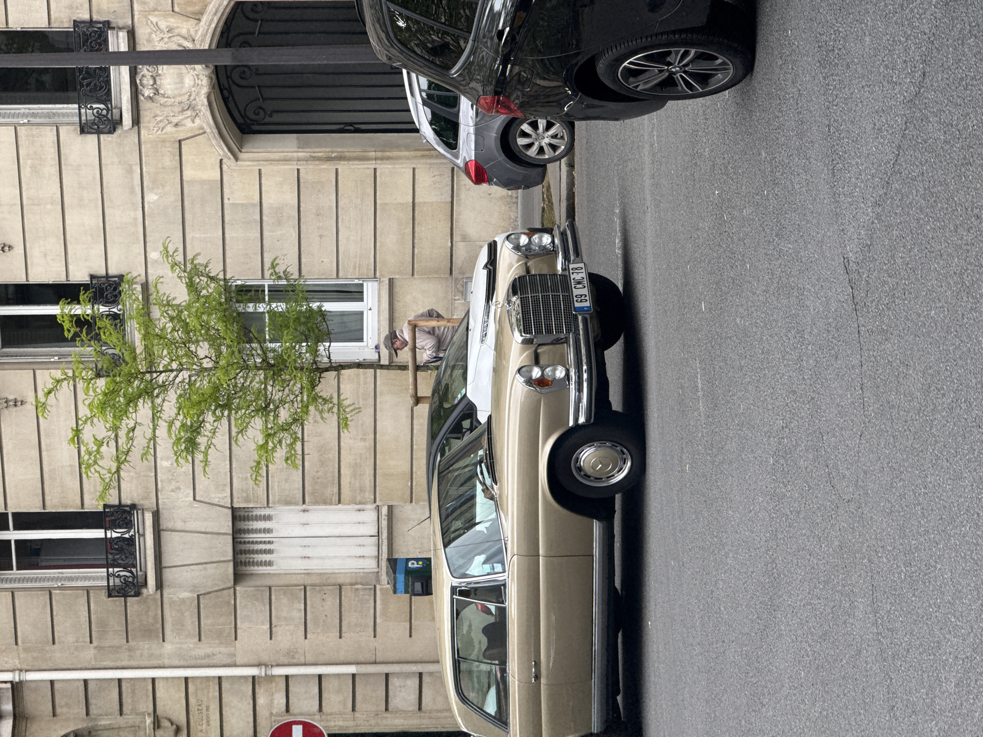 The image depicts a street scene in Paris, France, featuring a vintage, beige-colored Mercedes-Benz parked on the right side of the road. A black vehicle is parked closely behind it. A person wearing a light-colored shirt and hat appears to be working on or near the front of the Mercedes-Benz. The background consists of a Parisian building with distinct architectural features, including windows with black iron ornamentation and a stone facade. A small tree with green leaves is positioned near the building's facade. A street sign is barely visible in the bottom left corner. The street is paved with asphalt. The license plate on the Mercedes is visible and reads "69 CMC 18".