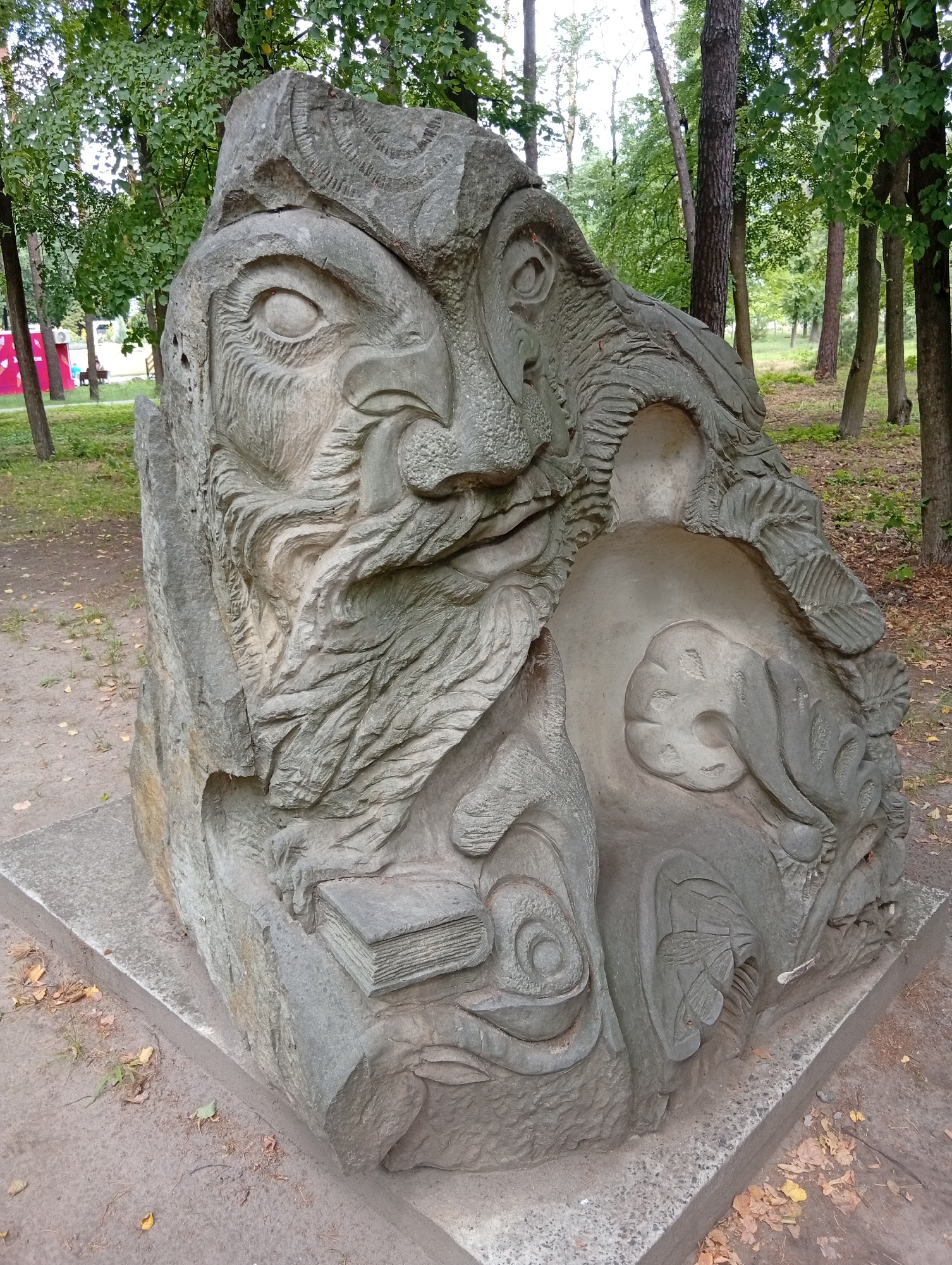 A stone sculpture of a face with a beard and an open book is displayed in a park. The sculpture is gray and appears to be carved from a single large rock. The face is depicted in profile, with a large eye and a prominent nose and beard. The beard is textured with swirling patterns. Below the face, a carved book lies open, with pages indicated by horizontal lines. The sculpture is situated on a concrete base in an outdoor environment with trees and foliage visible in the background. The ground around the sculpture is covered with dirt and fallen leaves. The lighting suggests it is daytime. The location is identified as Unknown, Unknown.