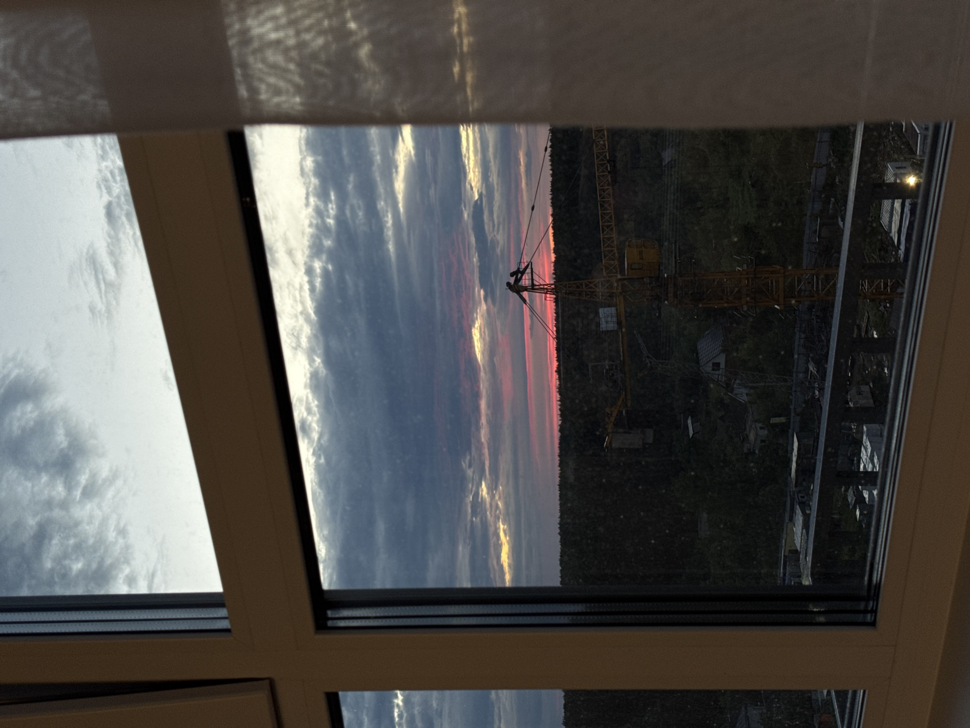 The image depicts a view from a window overlooking a landscape at dusk. The sky is filled with dramatic clouds, tinged with pink and yellow hues from the setting sun, suggesting it is either sunrise or sunset. A construction crane, yellow and imposing, stands prominently in the middle ground, its structure detailed with crisscrossing beams. The landscape below is dense with trees, forming a dark, forested area. In the distance, some buildings and structures, possibly part of a construction site or a small settlement, are visible amongst the trees. The foreground is framed by a white window frame, with a sheer white curtain to the right partially obscuring the view. The image appears to be taken from an elevated position, possibly an apartment building, in the city of Izhevsk, Russia.