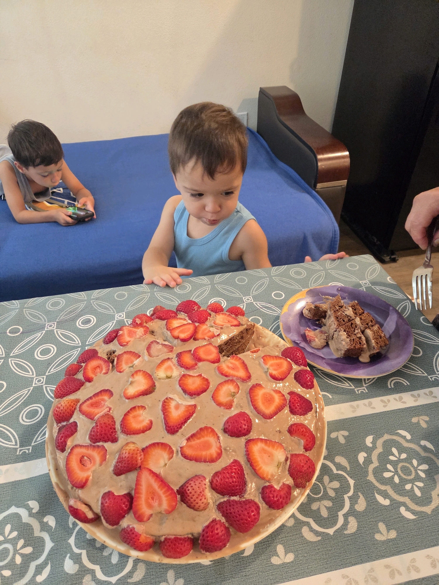 The image shows a birthday cake topped with strawberries on a table with a patterned tablecloth. Two young boys are present. One boy, wearing a blue tank top, is looking at the cake. The other boy, lying down and wearing a gray tank top, is playing on a handheld game device. A slice of cake is on a purple plate to the right of the cake, and a fork is held by a person whose hand is visible on the right side of the image. The background includes a blue sofa and a dark piece of furniture. The scene appears to be indoors, likely in a residential setting.