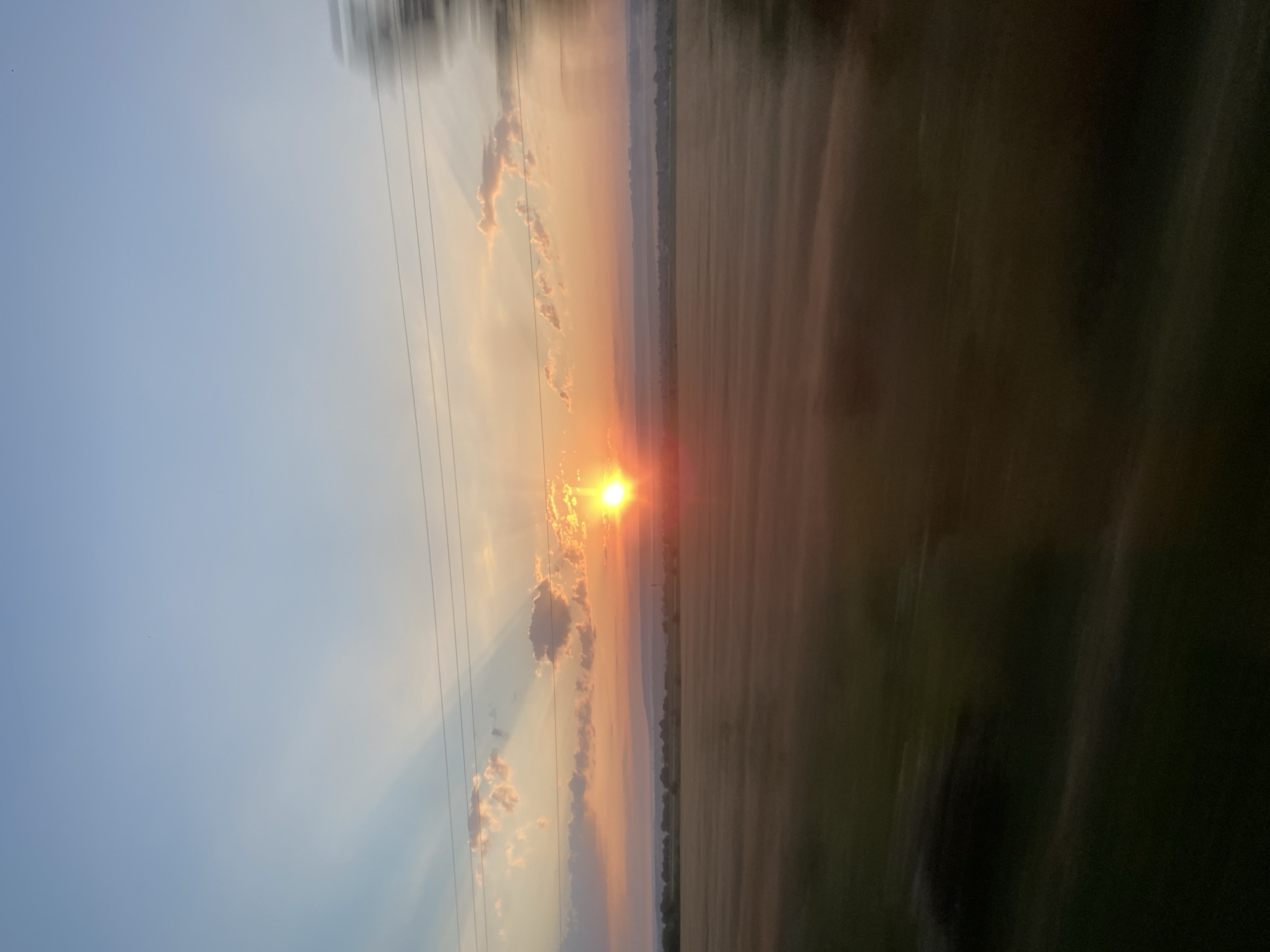 A sunset viewed from a moving vehicle in Novosiolovca, Moldova, casts a warm glow across a rural landscape. The sun, a bright orange orb, is positioned slightly to the left of the frame, its rays fanning out through wispy clouds. The sky transitions from a clear, pale blue at the top to a soft orange and pink hue near the horizon. Three thin power lines stretch horizontally across the upper portion of the image. Below the sky is a vast field, likely agricultural, with a hazy or misty quality. In the far distance, a line of trees and subtle undulations suggest the topography of the land. The motion blur indicates the photograph was taken from a moving car, with the right side of the frame showing more pronounced streaking, especially on the blurred green and dark foliage.