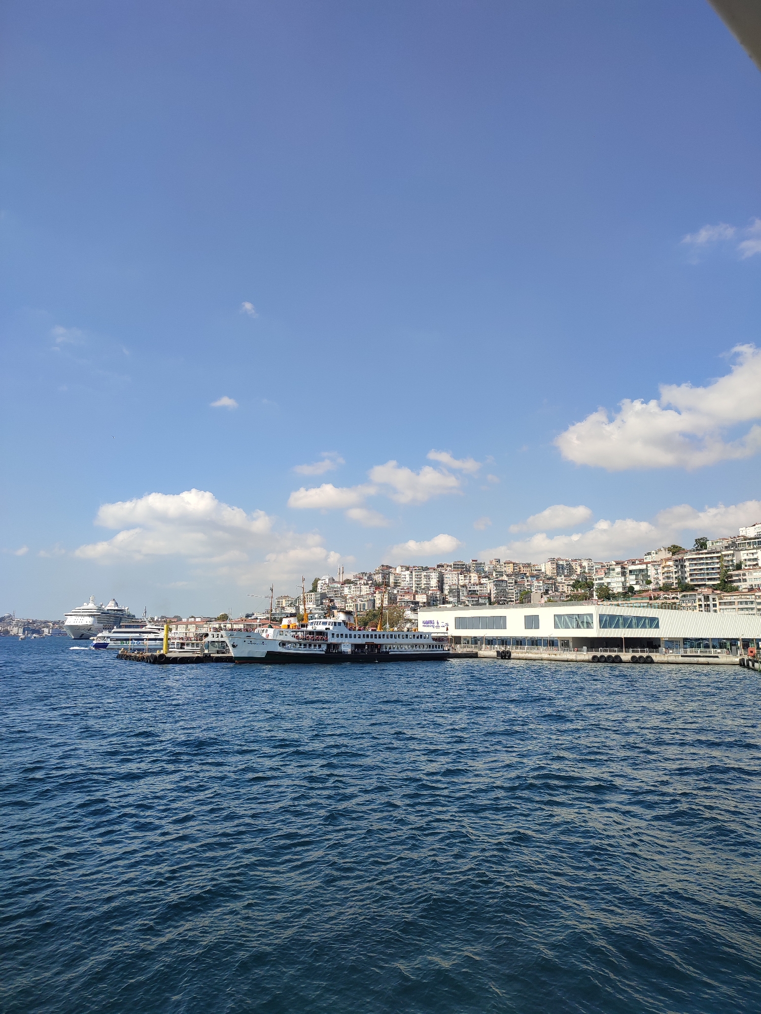 The image depicts a wide shot of a waterfront with several ferries docked at a pier in front of a densely populated city skyline. The water is a deep blue with visible ripples. A large, modern white building with numerous windows forms the waterfront structure, with a long pier extending outwards. Several white and green ferries are moored along the pier. In the distance, to the left, a large white cruise ship is visible. The city skyline behind the pier is characterized by a dense cluster of buildings, many with white facades and red tile roofs, cascading up a hill. A tall, slender minaret is visible above the buildings on the far left, suggesting a mosque. The sky is a clear, light blue with scattered white clouds. The overall impression is that of a bustling urban port. The view is consistent with the Kabataş ferry terminal in Beyoğlu, Istanbul, Turkey, a major transportation hub connecting the European side of Istanbul to its Asian side.