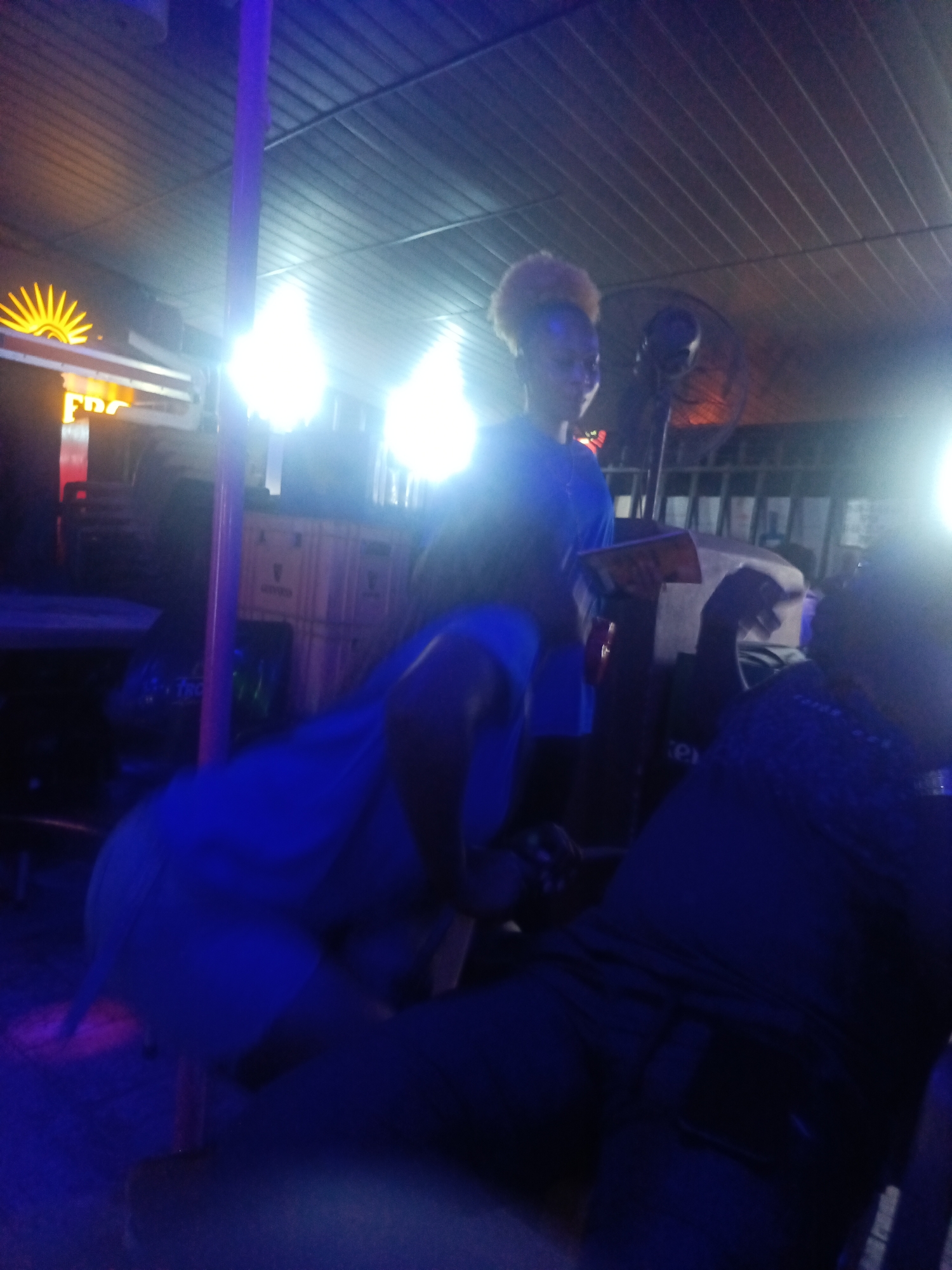 In the city of Unknown, Unknown, at night, a woman with blonde, curly hair is seen posing near a pole. She is wearing a blue outfit. In the background, a large ceiling fan is visible, and there are bright lights illuminating the scene, casting a blue hue over everything. Another person, wearing a dark blue top, is also present, though their face is not clearly visible. The environment appears to be an indoor venue, possibly a bar or club, with a corrugated metal ceiling. There are also some crates and tables visible, suggesting a commercial or entertainment setting. The overall atmosphere is dimly lit, with a strong emphasis on artificial lighting.
