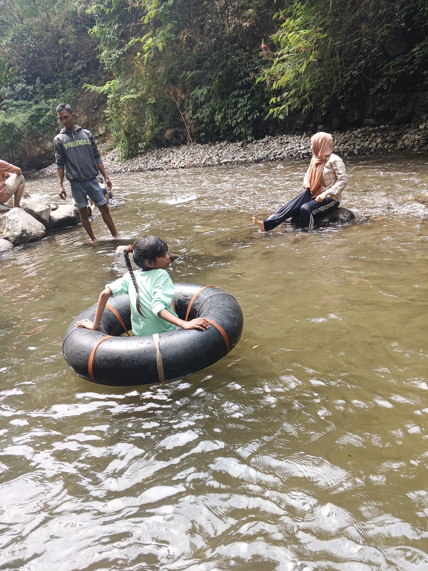 In Pacet, Indonesia, a group of individuals is enjoying a day by a river. In the foreground, a young girl with a braided ponytail is seated in a black inner tube, wearing a light green t-shirt. She is facing towards the right of the frame, her arms resting on the tube. To the left, a man in a gray hoodie and denim shorts stands in the shallow river, looking downwards. Further back, a woman wearing a headscarf and a checkered long-sleeved shirt is seated on a rock in the river, with her legs extended. The riverbed is lined with numerous stones and pebbles, and lush green vegetation covers the steep embankment in the background. The water is murky brown, reflecting the sunlight and creating patterns on its surface.