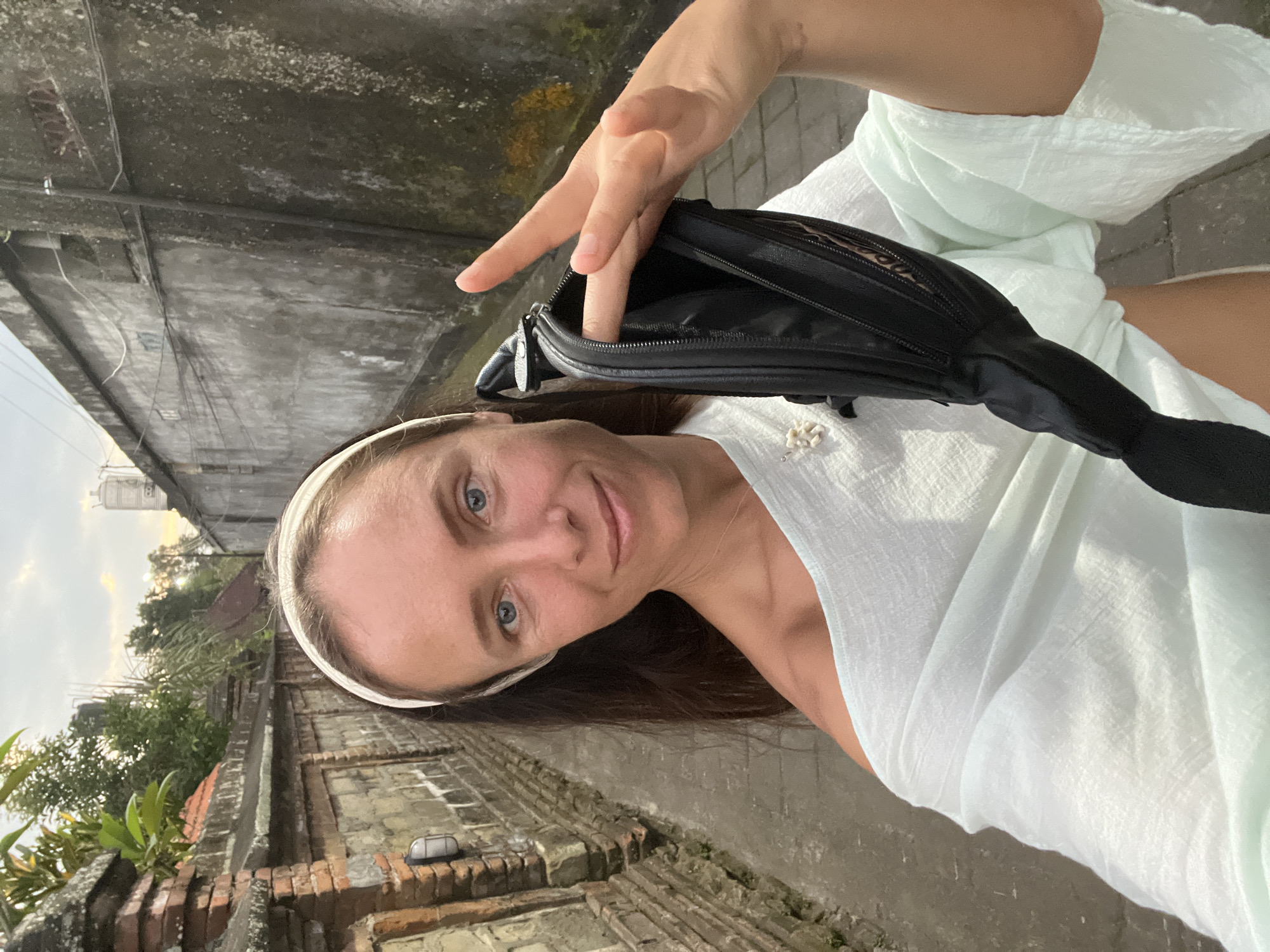 In a narrow alleyway in Ubud District, Indonesia, a woman with blue eyes and long brown hair wears a white headband and a light blue off-the-shoulder top. She is holding a black fanny pack, with her finger inside the open pouch, and making a peace sign with her other hand. The alley walls are made of brick and concrete, and there is vegetation and a water tank visible in the background. The ground is paved with cobblestones. The scene appears to be outdoors, possibly during the day.