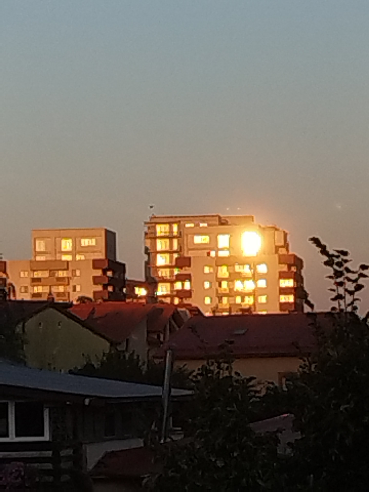 The image shows a cityscape with apartment buildings and houses bathed in the warm glow of sunset. The sun is prominently visible, reflecting brightly off the windows of a tall apartment building, creating a luminous effect. The sky is a gradient of soft orange and pale blue, typical of twilight. In the foreground, rooftops of houses and some greenery are visible, suggesting a residential area. The architecture appears to be typical of Eastern European cities.