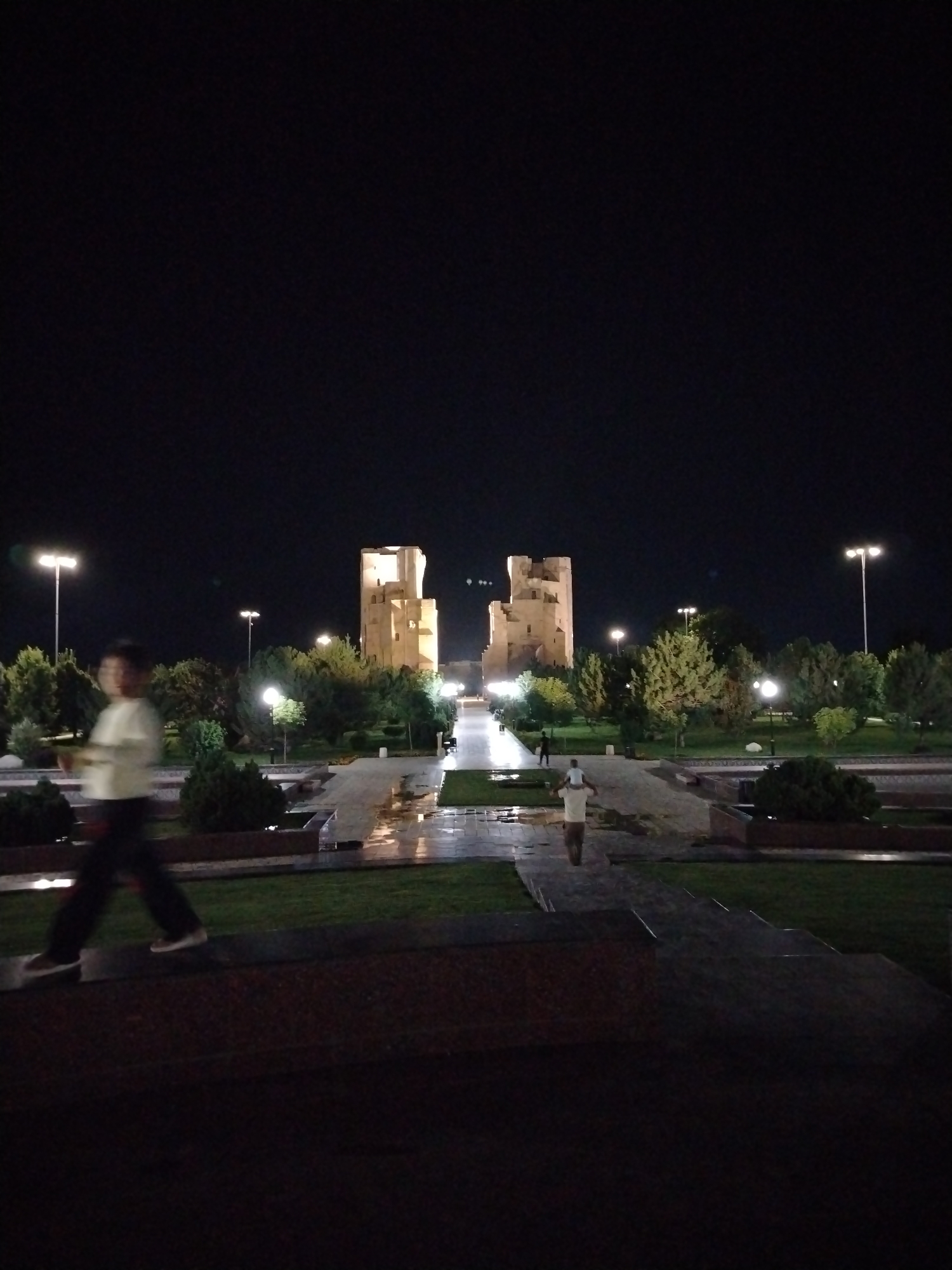 The image depicts a night scene in Shahrisabz City, Uzbekistan, featuring the Ak-Saray Palace ruins. The ruins, illuminated by artificial lighting, are the central focus in the mid-ground. In the foreground, to the left, a person is captured in motion, appearing as a blurred figure walking from left to right. To the right, a second person is visible, also in motion, facing away from the camera and walking towards the ruins. The environment includes manicured lawns, paved walkways that appear wet and reflective, and trees. Streetlights are visible in the background, illuminating the area and contributing to the overall nighttime ambiance. The sky is dark, indicating it is night.