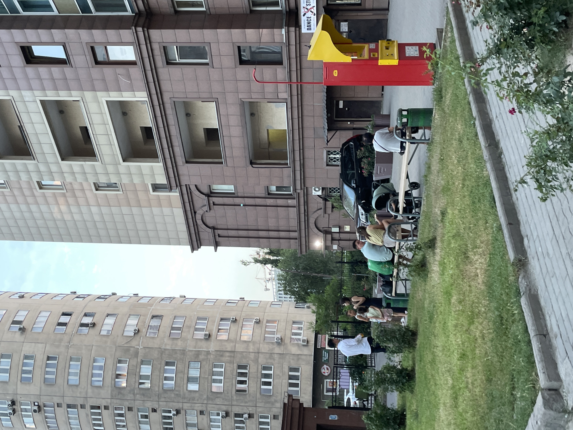 A street scene in Almaty, Kazakhstan, captures a group of people gathered on a bench in a grassy area, with modern buildings in the background. A tall, curved residential building with numerous windows and air conditioning units stands to the left, while a more angular, beige-and-brown building with balconies is to the right. Several individuals, including adults and children, are seated and standing near a picnic bench. A black SUV is parked in front of the right building, and a bright red and yellow automated payment kiosk is positioned near its entrance, adjacent to a sign for "DANCE STUDIO". The overall environment suggests a residential urban setting.