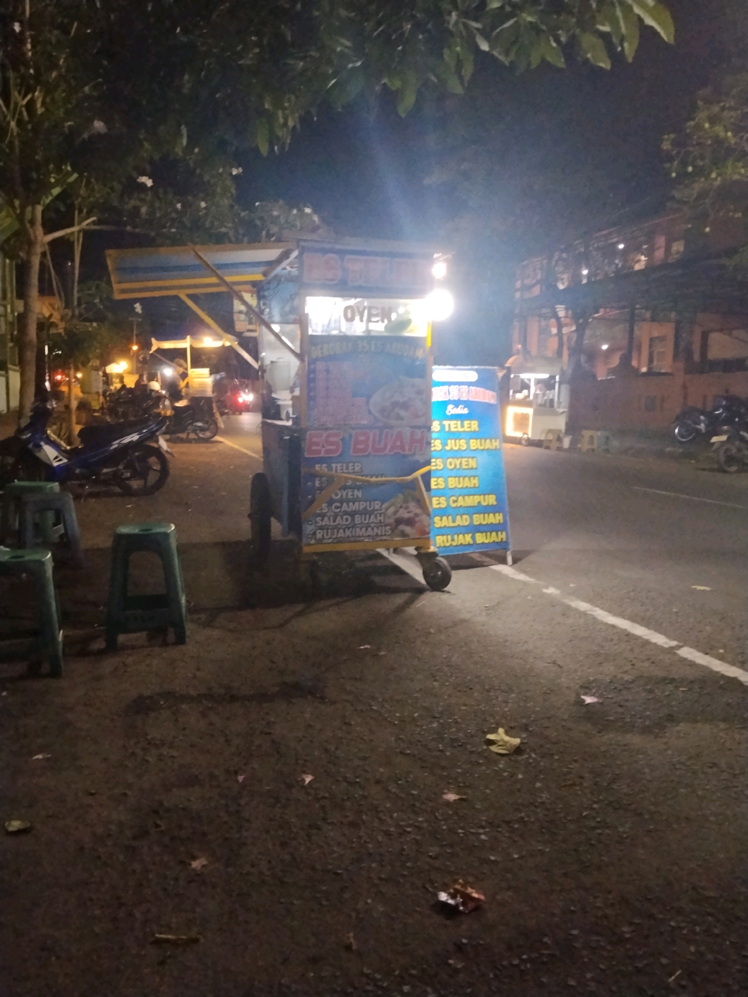 This is a nighttime, eye-level shot of a mobile food cart selling various fruit-based drinks and desserts. The cart is blue and yellow, with signage in Indonesian listing items like "Es Teler," "Es Jus Buah," "Es Oyen," "Es Buah," "Es Campur," "Salad Buah," and "Rujak Buah." The cart is positioned on the side of a paved road, likely in an urban setting given the presence of buildings and streetlights in the background. Several green plastic stools are placed in front of the cart, suggesting a waiting or seating area for customers. A blue and white motorcycle is parked to the left of the cart. The road appears wet, possibly from recent rain or street cleaning. The background shows buildings with illuminated windows and more streetlights, indicating a commercial or residential area. The overall atmosphere is dimly lit, with the food cart being a prominent source of light.