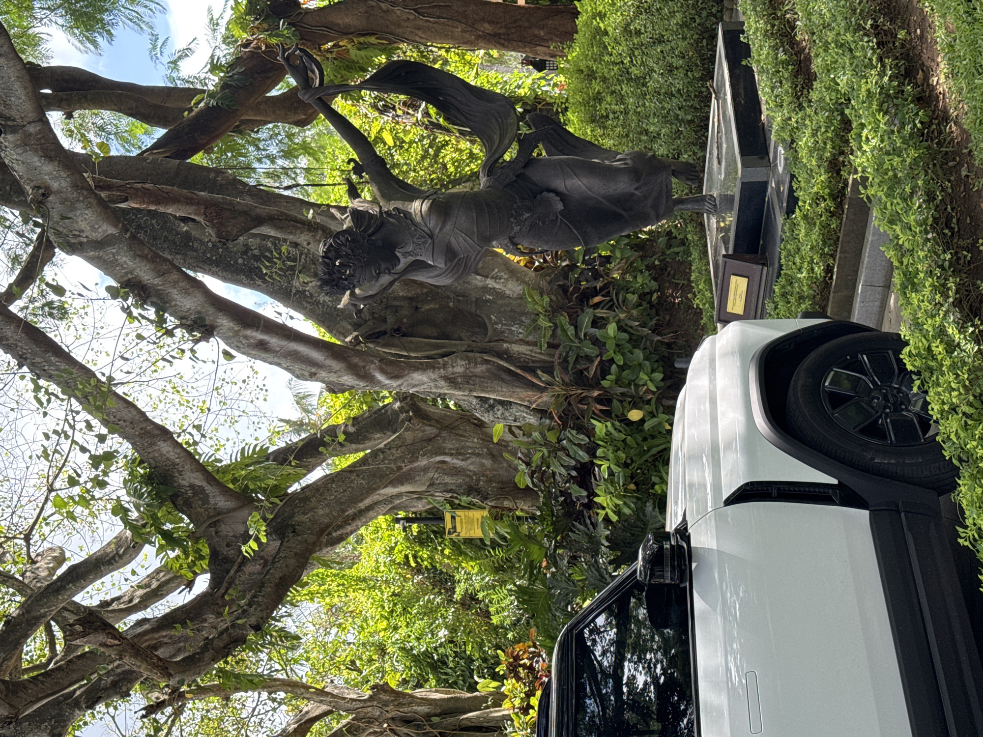 The image shows a statue of a woman holding a scarf, likely located in Kuta Selatan, Indonesia. The statue is positioned beneath a large, mature tree with exposed roots and dense foliage. A white vehicle is partially visible on the right side of the image, suggesting the statue is located near a road or parking area. The background includes dense greenery and a glimpse of the sky. The statue sits on a base that looks like a grave stone.