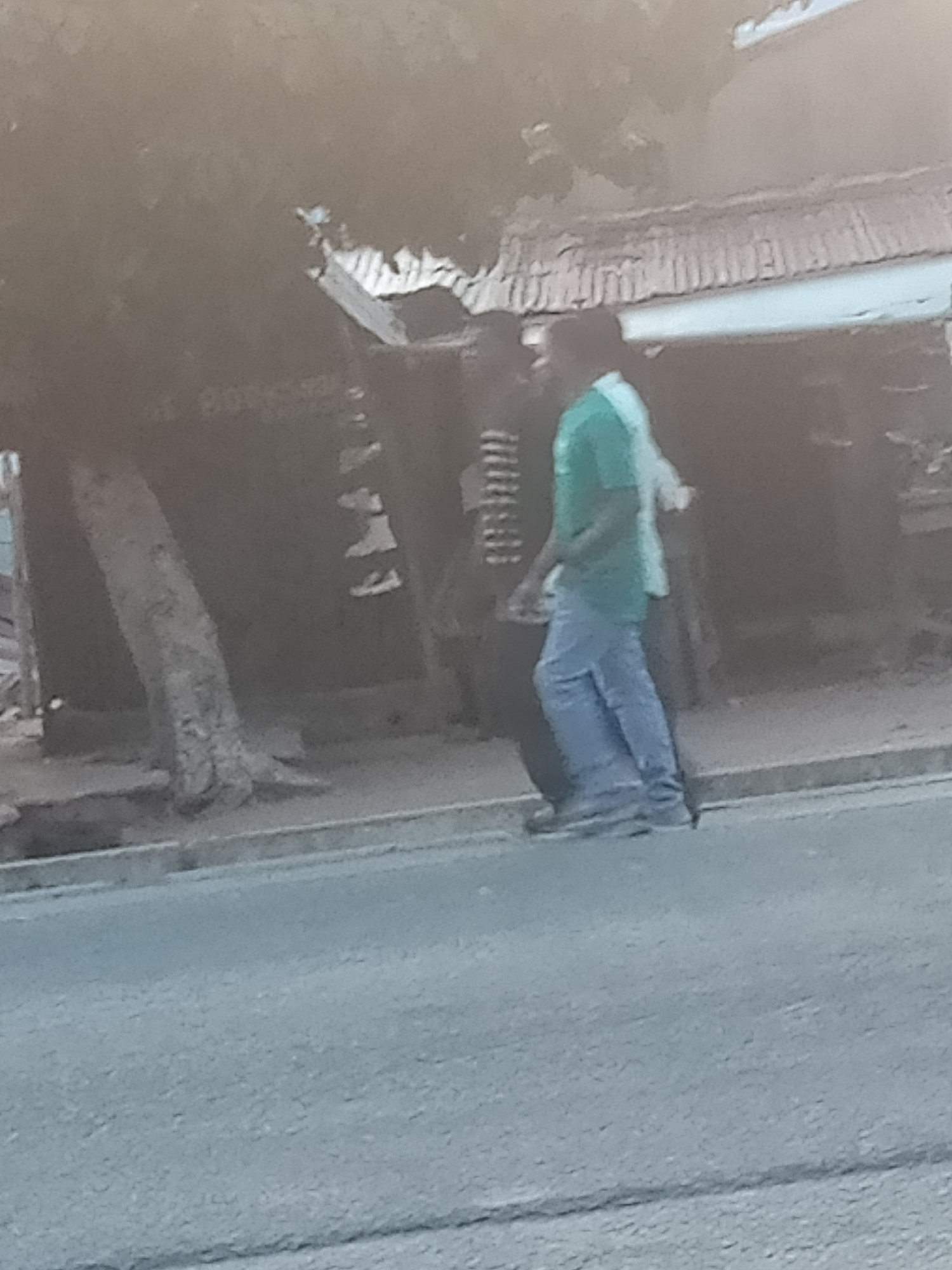 Two individuals, one wearing a green t-shirt and blue jeans, and another person whose attire is not clearly discernible, are walking along a paved road. The person in the green t-shirt is in the foreground and appears to be walking towards the right of the image, with their left leg extended. Behind them, the other individual is partially obscured. The background features a building with a corrugated roof and a tree trunk. The scene is outdoors, with a street in the foreground. The image appears to be taken in Jalingo, Nigeria.