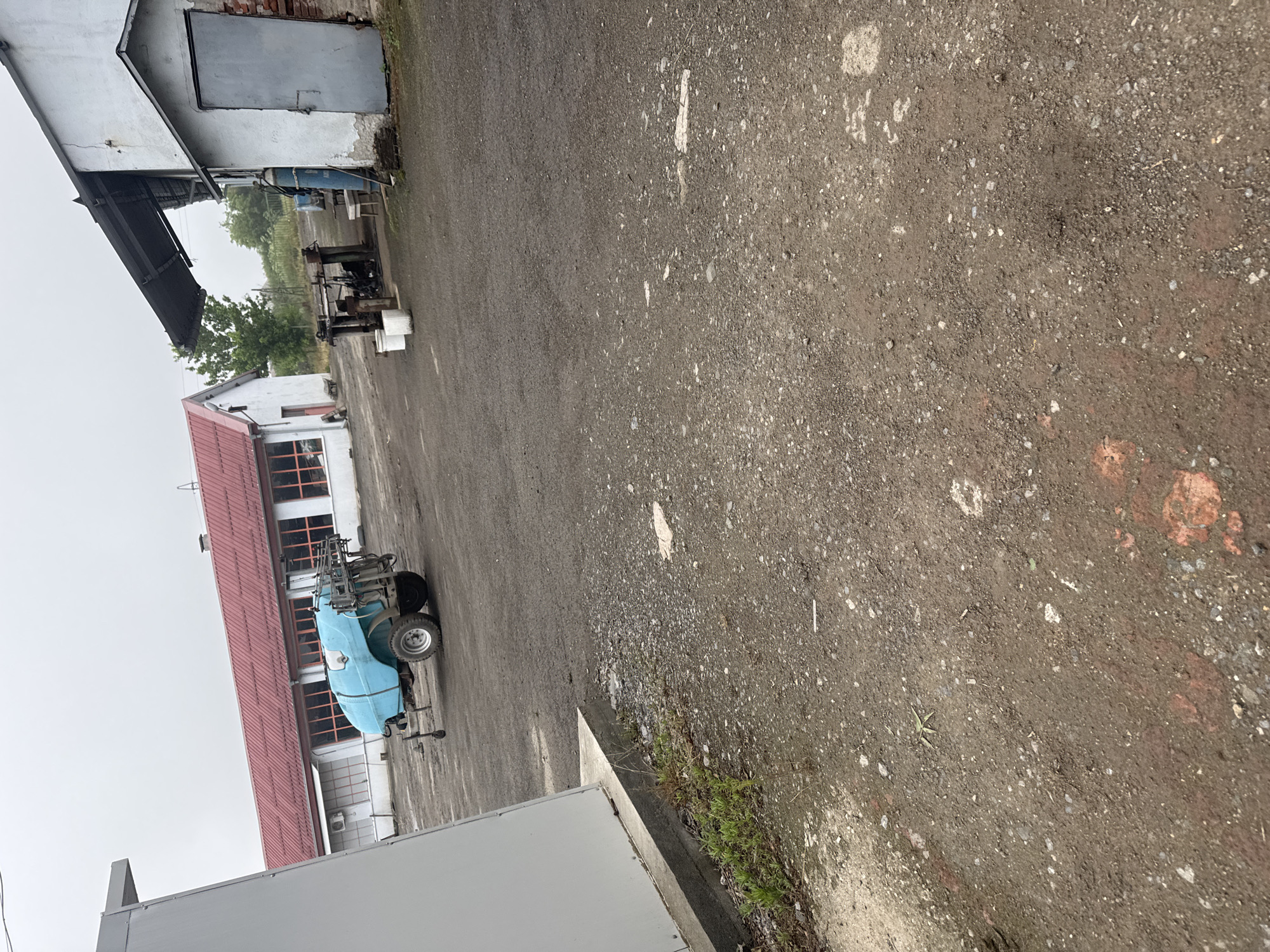 The image captures an outdoor yard or lot, likely part of an agricultural or industrial facility, under an overcast sky. The ground is a mixture of dark gravel and dirt, exhibiting varying textures and lighter patches.
In the mid-ground, a light blue agricultural sprayer with wheels, possibly a field or orchard sprayer, is prominently parked in front of a white building with a red roof and multiple windows. Further back on the left, another white building features a dark, corrugated metal overhang or shed structure. Adjacent to this building, several pieces of dark, older-looking machinery, resembling parts of farm equipment, are visible, alongside white buckets or containers. A metal door is noticeable on one of the buildings. The foreground shows the corner of a light-colored building or structure, with green vegetation, including grass and weeds, growing along its base.
The overall scene suggests a functional outdoor storage or work area. The sky is uniformly gray, indicating a cloudy day with diffused lighting and no direct sunlight. No people are present, and no specific activity is taking place. There is no visible text. The setting is consistent with an agricultural environment, aligning with the provided location of Sarnevo, Bulgaria.