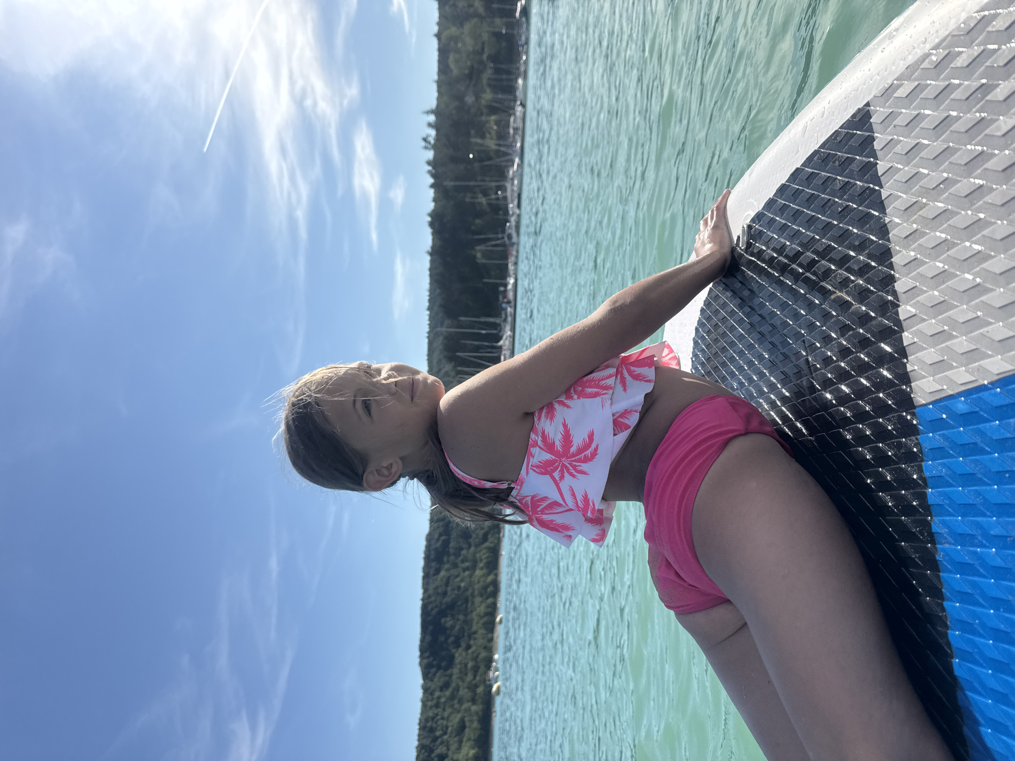 A young girl in a pink palm-print bikini is lying on a paddleboard in the water. She is looking up at the sky with her hands on the paddleboard. The sun is shining, and there are clouds in the sky. The water is green and calm, and there are trees in the background. The girl appears to be relaxed and enjoying herself. The image is taken from a slightly low angle, looking up at the girl and the sky. The paddleboard has a blue and gray textured surface.