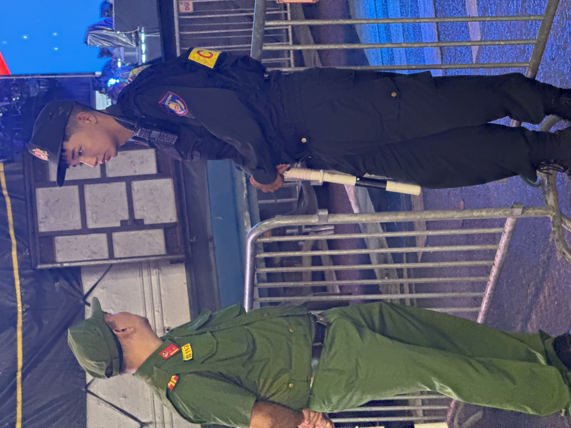 Two men in uniform stand on a street at night in Hanoi, Vietnam, behind a silver metal barricade. The man on the left wears a green military uniform with a green cap, epaulets with red and yellow insignia, and a yellow badge with red text. He has a dark belt and green pants. The man on the right wears a dark uniform with a dark cap, a radio on his chest, and a patch with a circular emblem and the word "CO DONG". He has a yellow band on his left arm with the letter "C". He holds a white baton with black stripes. Behind them is a dark structure with square panels, and a large blue screen is visible in the background. The ground appears wet, suggesting recent rain.