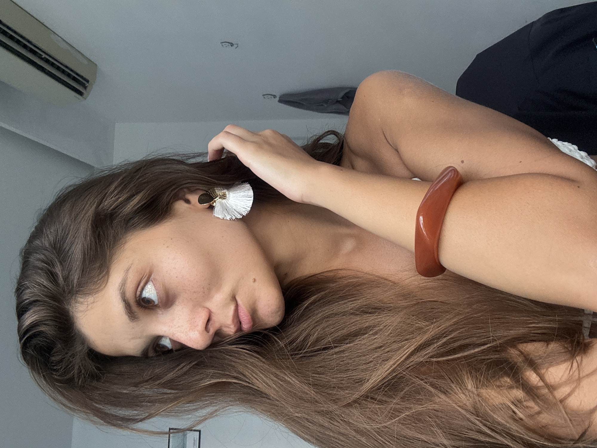 A woman with long, brown hair and freckles lies sideways on a surface. She is wearing a white tassel earring and a brown bangle on her arm. Her eyes are partially open. She is touching her hair with her hand. The background includes a white ceiling with an air conditioning unit and a dark object. There is no indication of this location being in the city of Đà Nẵng, Vietnam.