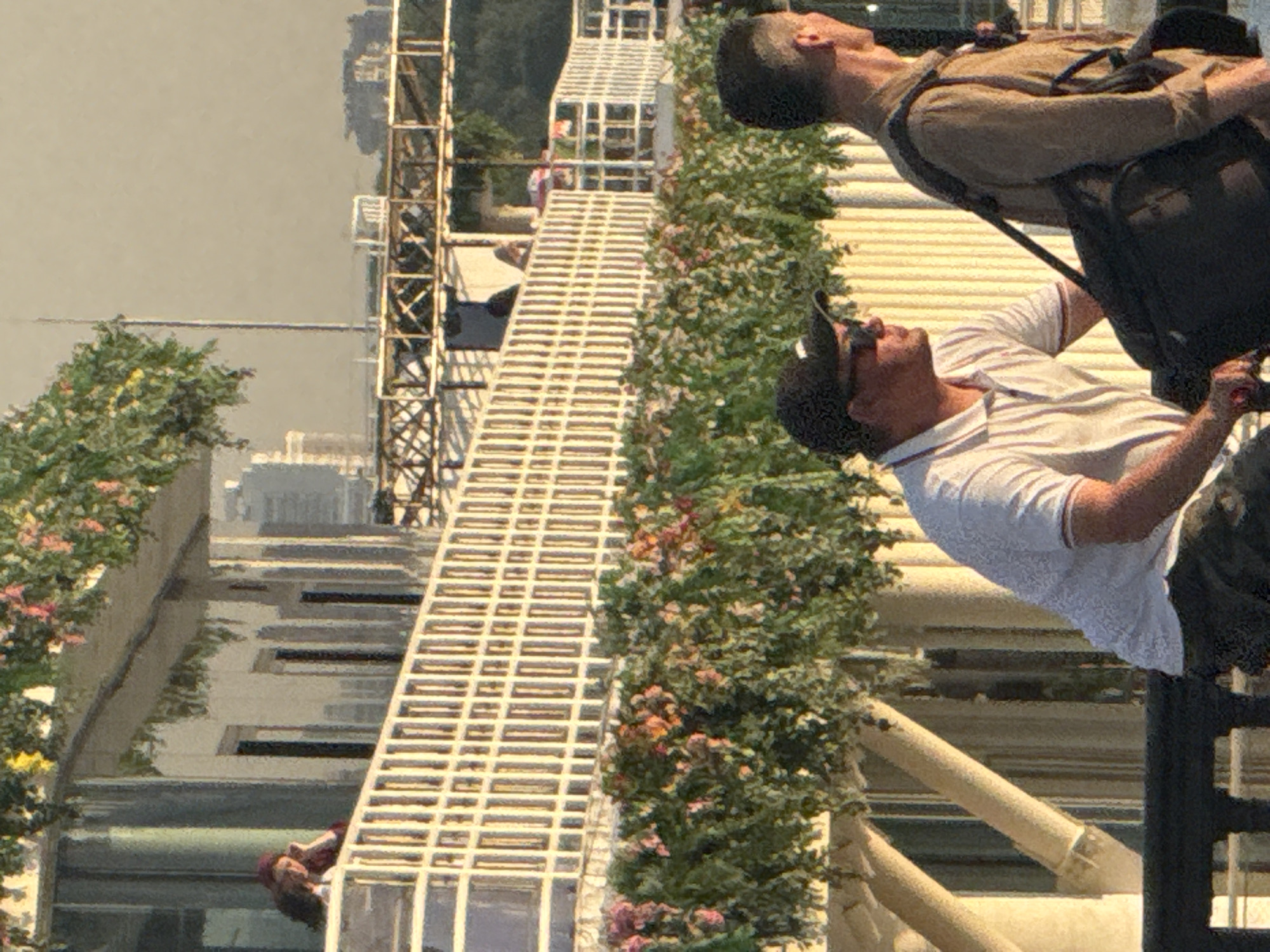 In the city of Unknown, Unknown, two men are visible in the foreground of the image. The man on the left wears a white collared shirt, a black baseball cap, and sunglasses. He is looking to his right. The man on the right wears a brown collared shirt and has a backpack slung over his right shoulder. He is looking to his right, slightly away from the other man. Both men appear to be walking or standing on a raised platform.
In the background, a building with a white railing and hanging plants is visible. There are several people on a balcony on the building. The sky is a hazy, light color. The scene appears to be outdoors during daylight.