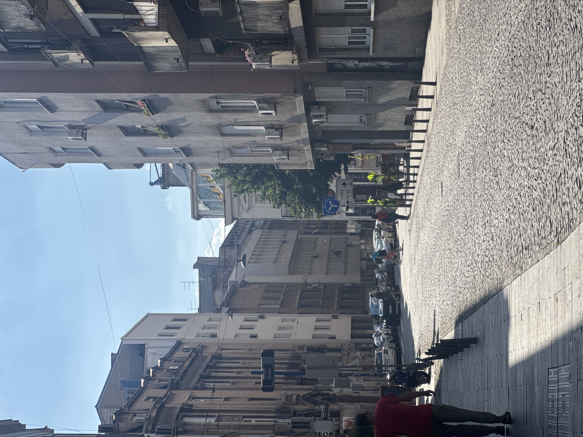 The image depicts a sunny day on a cobblestone street in Belgrade, Serbia. In the foreground, a man wearing a red shirt and khaki pants walks away from the camera down a tiled sidewalk. The street is lined with multi-story buildings constructed in a mix of architectural styles, some appearing older and ornate, while others are more modern and utilitarian, featuring balconies and air conditioning units. Cars are parked along the right side of the street, and several people are walking or crossing the street. A traffic sign with Cyrillic lettering is visible on the left side of the street. The atmosphere suggests a typical urban scene in a European city.