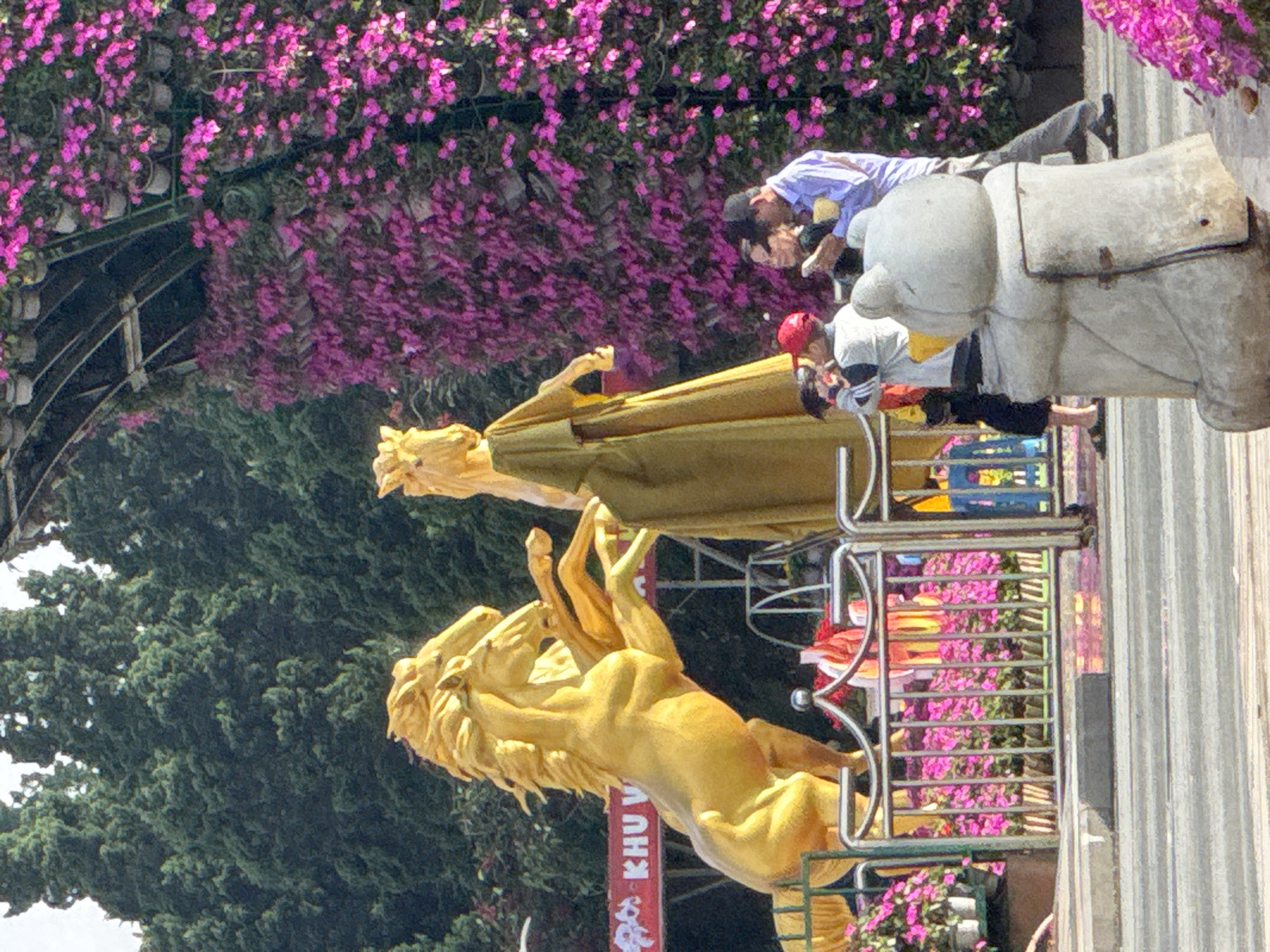 The image depicts a public space in Phường Xuân Hương - Đà Lạt, Vietnam, featuring large golden horse statues. In the foreground, a white, bear-like statue is visible. Behind the statues, a vibrant vertical garden of pink flowers cascades down a structure. Several people are present, including a man in a blue shirt and a black cap, a woman in dark clothing, and a person in a grey jacket with a red cap. One person is holding a phone. In the background, lush green trees form a dense forest. A red banner with white text is partially visible. The ground appears to be paved with a distinct curb and road surface.