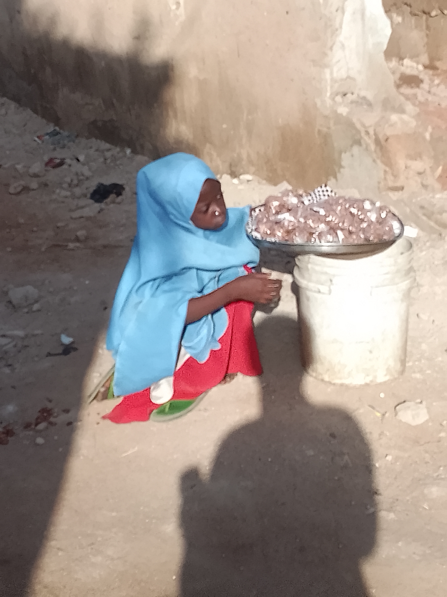 A person wearing a blue headscarf and a red garment sits on the ground in Pantame, Nigeria, with a shallow metal dish filled with small food items on their lap. The food appears to be a mixture of grains and possibly pieces of meat. The person's left hand is visible, reaching towards the dish. To the right of the person is a tall, white plastic bucket. The background consists of a rough, textured wall, likely made of mud or concrete, casting a shadow on the ground. The ground is covered in dirt and small pebbles, with some scattered debris. The overall scene suggests an outdoor environment, possibly a market or a residential area.