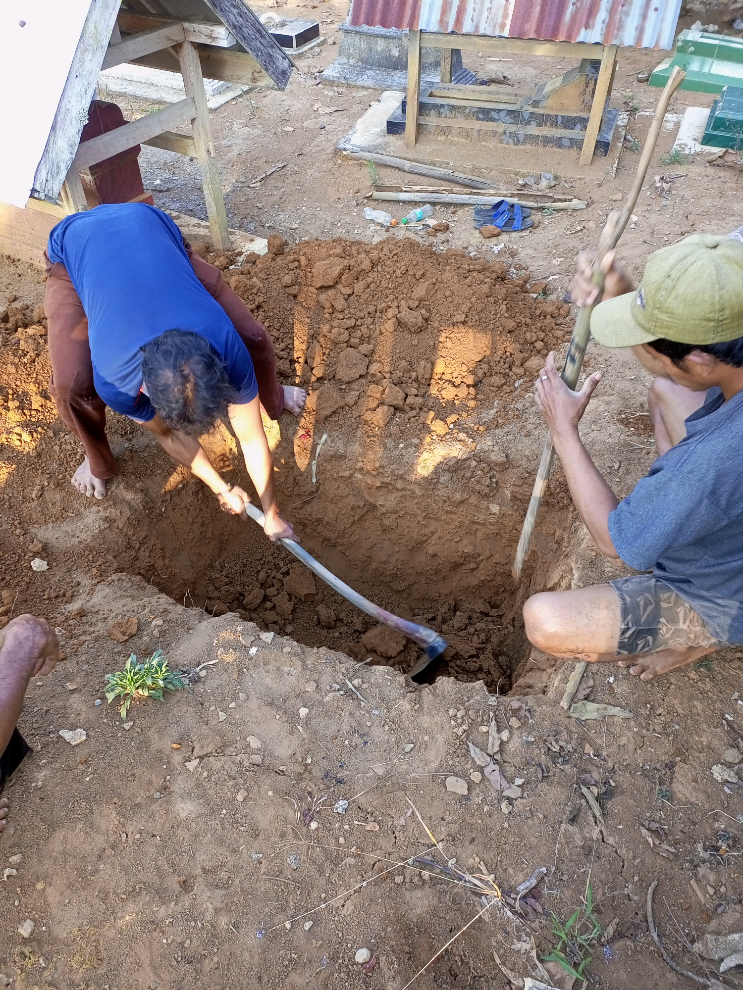 In Jatipurno, Indonesia, two individuals are engaged in digging a grave. The scene depicts a cemetery with gravesites visible in the background, some with rudimentary shelters or markers. The foreground is dominated by a freshly dug, rectangular grave. One person, wearing a blue t-shirt and brown pants, is actively digging using a shovel. The other individual, wearing a green cap and a gray t-shirt, is also using a shovel, positioned to the right of the grave. The ground is composed of brown soil, with some dirt and debris scattered around the excavation. The overall atmosphere is one of manual labor and preparation for a burial.