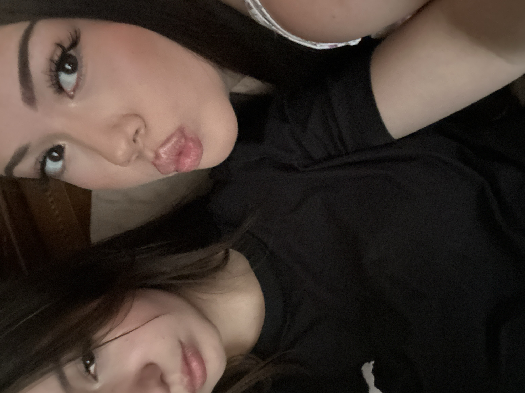 A tilted, close-up selfie captures two young women, likely indoors in Naberezhnye Chelny, Russia. The woman in the foreground is prominent, facing the camera with her head tilted to the side. She has dark hair, well-defined eyebrows, and long eyelashes, with light blush on her cheeks. Her lips are puckered in a playful "kissing" pose as she looks upwards. She is wearing a dark top, with a light-colored, possibly patterned, strap visible near her upper chest.
Partially visible below and behind her is a second woman, also with dark hair that falls slightly over her forehead. She is also looking upwards, with a more subtle, relaxed expression and slightly parted lips. She appears to be wearing a dark, possibly black, top.
The background is largely dark and out of focus, offering no distinct details other than a hint of lighter, possibly upholstered, material behind the second woman. The image conveys a casual and friendly moment between the two individuals. There are no visible signs of specific weather or time of day, and no text is present.