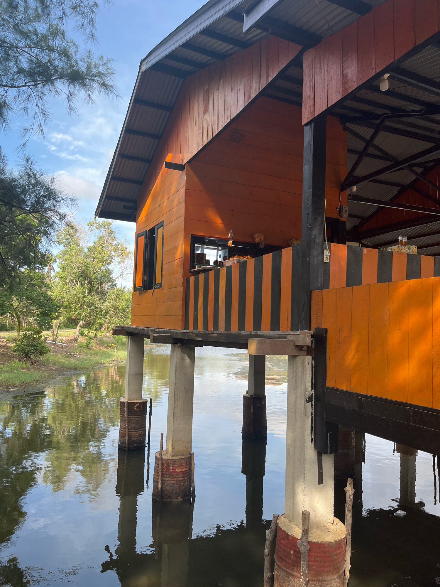 An elevated wooden building with an orange facade and a corrugated metal roof stands on concrete pillars over a body of water. The building has a dark band of orange and black striped wood around its lower section, and appears to be a structure in a rural or natural setting. The water reflects the building and the surrounding trees and sky. The scene is located in Aneuk Paya, Indonesia.
