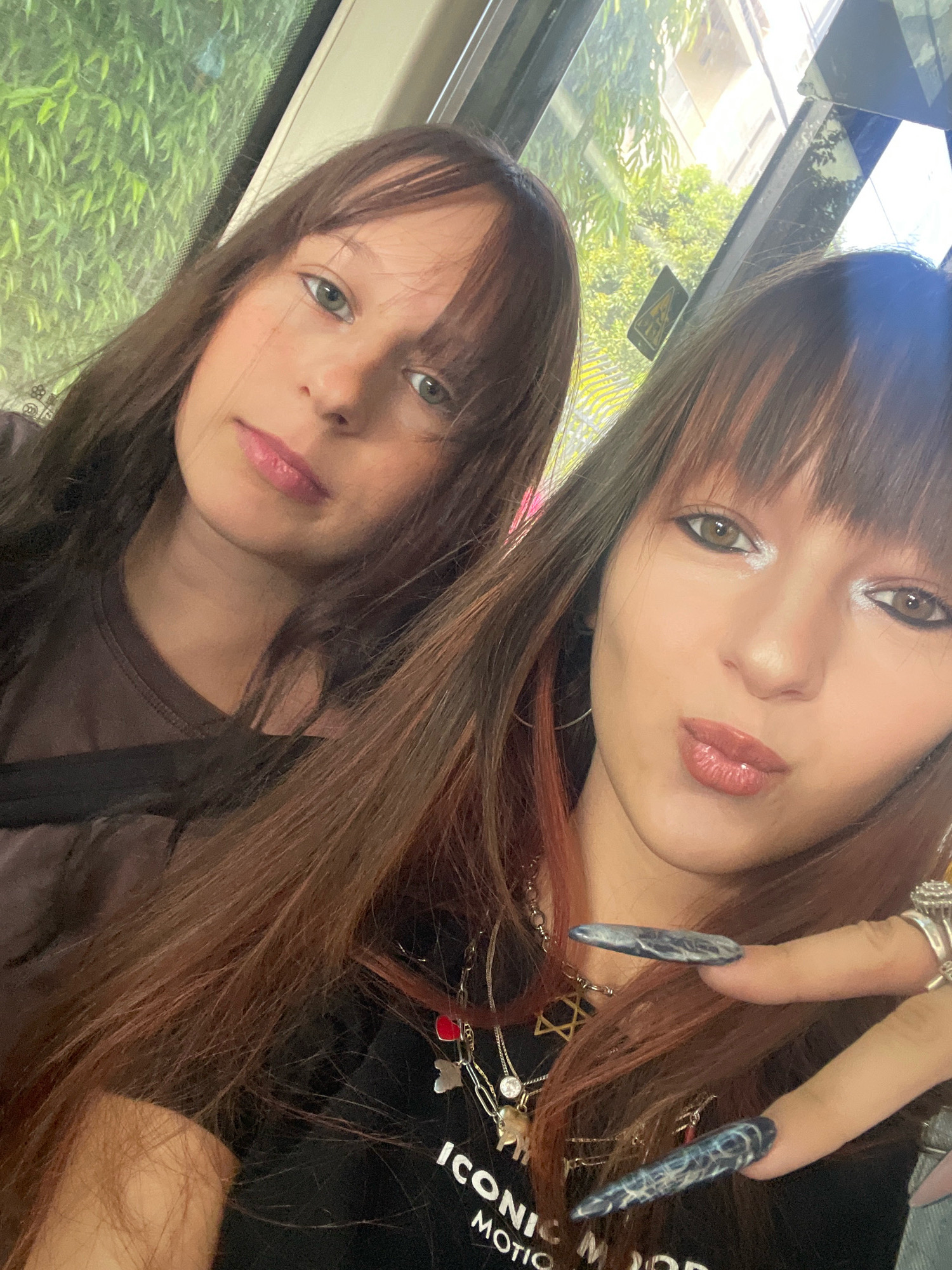 Two young women are taking a selfie, likely inside a vehicle. The woman on the right is in the foreground, with her face angled towards the camera. She has long, brown hair with reddish highlights and bangs. Her makeup includes dramatic eyeliner and shimmering eyeshadow. She is wearing a black t-shirt with a silver necklace featuring multiple charms, including a Star of David. She is also wearing several rings on her fingers, one of which is a large silver ring. Her fingernails are long and painted with a dark, swirled design. The woman on the left is slightly behind and to the left of the other woman. She also has long, brown hair with bangs and is wearing a brown t-shirt. Both women appear to be looking at the camera. The background suggests they are in a moving vehicle, with a window showing greenery and buildings outside, indicating an urban or suburban environment. The sunlight filtering through the window suggests it is daytime. The presence of a yellow triangular warning sign on the window may indicate they are on public transportation. Given the specific mention of Raanana, Israel, this scene could be taking place on a bus or a private vehicle traveling through the city.