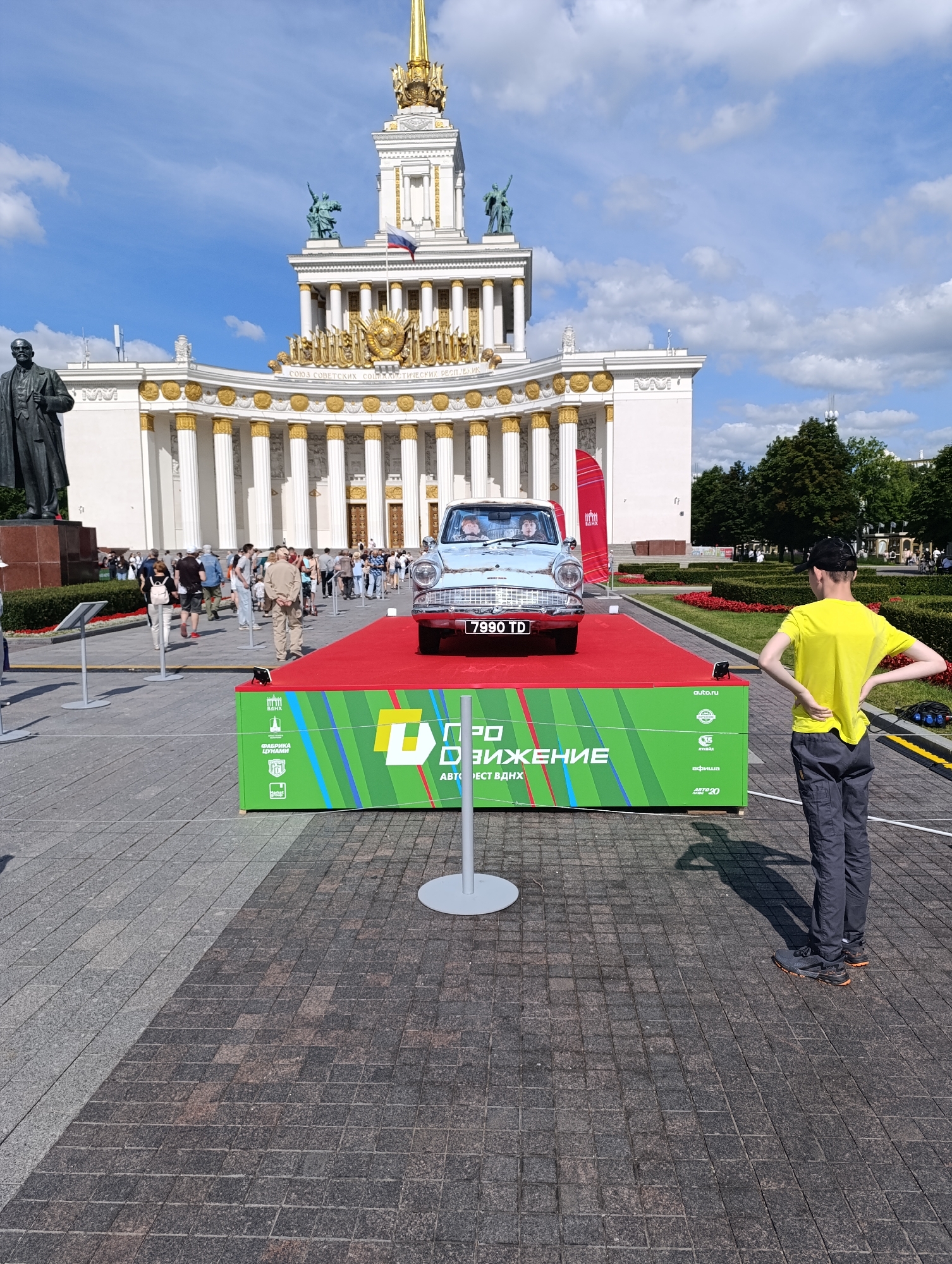This image captures an outdoor scene in front of a grand, white building with gold embellishments, which is a prominent landmark in Moscow, Russia, specifically at the VDNKh (Exhibition of Achievements of National Economy). In the foreground, a light blue vintage car, likely a Ford Anglia, is displayed on a red carpeted platform. The car has the license plate "7990 TD". The platform is supported by a green box with colorful stripes and text in Russian, including "Движение" (Movement) and logos. To the right of the platform, a young person in a yellow t-shirt and dark pants stands with their hands on their hips, facing the car. Behind the platform, a crowd of people are walking and observing. To the left of the car, a large bronze statue of a man in a suit stands on a pedestal. The background shows the impressive architecture of the VDNKh pavilion, adorned with columns and statues, under a bright blue sky with scattered clouds. The ground is paved with dark gray square tiles, with a lighter gray paved area to the left. Green trees and bushes are visible in the distance, bordering the paved area. The overall atmosphere appears to be a public event or exhibition on a sunny day.
