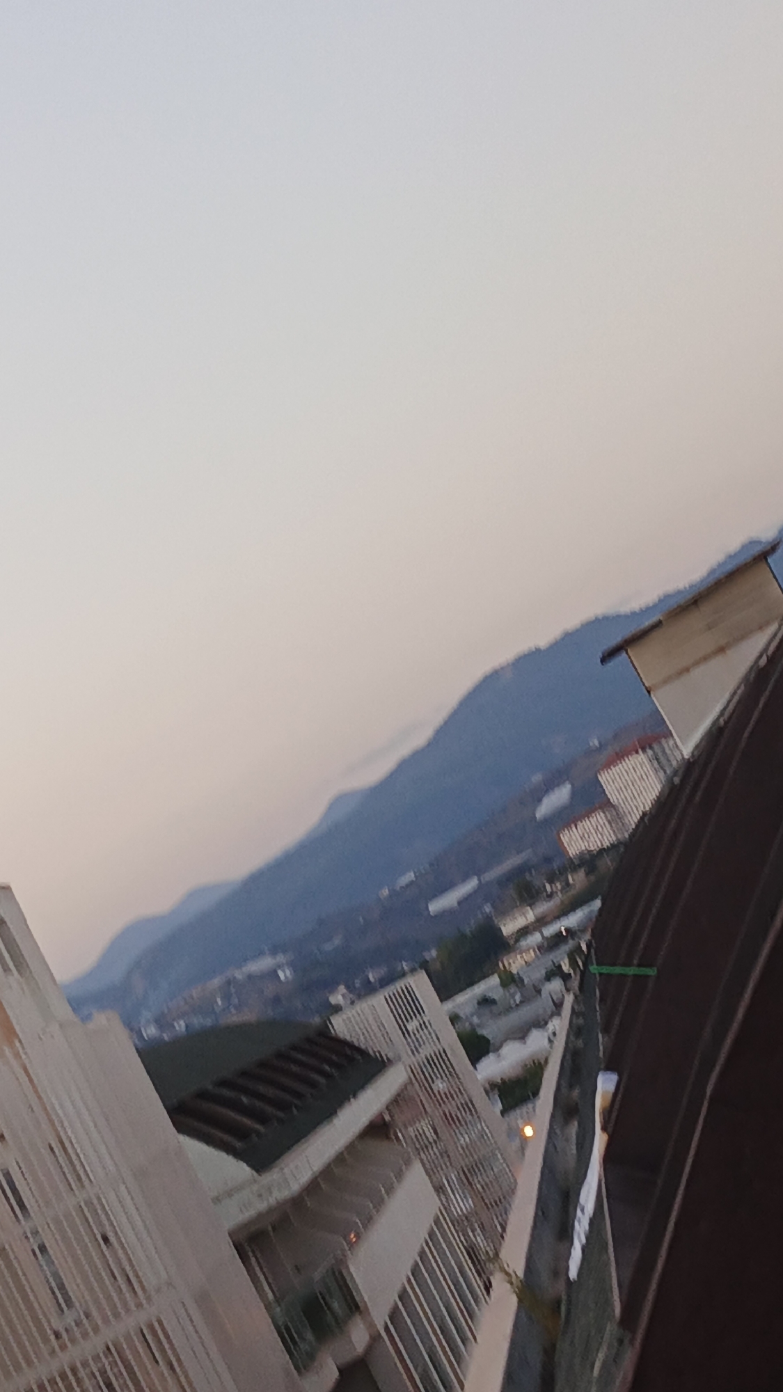 The image is an outdoor, slightly tilted, medium-angle shot capturing a cityscape at dusk. The sky is a pale, hazy gradient of white and light gray, suggesting twilight. In the midground, a range of blue-tinted mountains forms the horizon, with some scattered buildings visible on their slopes and in the valley below. In the foreground, two modern buildings are prominent. The one on the left is a light-colored, multi-story structure with a white, latticed facade. The building on the right is partially obscured by a dark brown, corrugated material, possibly a roof or wall, that dominates the right side of the frame. White vertical railings are visible along balconies of both buildings, and some windows are present. A few sparse green plants can be seen on the buildings' ledges and balconies. The overall impression is of a residential or commercial area in Alanya, Turkey, looking towards the mountainous landscape. There are no people or visible actions in the scene.