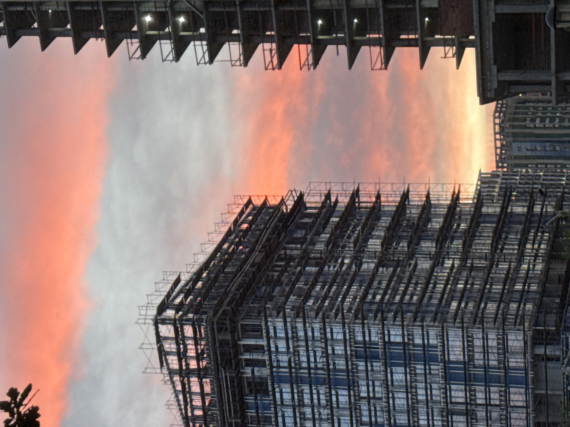 The image shows a low-angle view of unfinished buildings against a striking sunset sky. The buildings are steel frame structures, typical of construction sites. The sky transitions from orange and pink hues near the horizon to gray and white towards the upper parts of the frame. The photograph is rotated. A hint suggests that the photo might be located in Izhevsk, Russia.