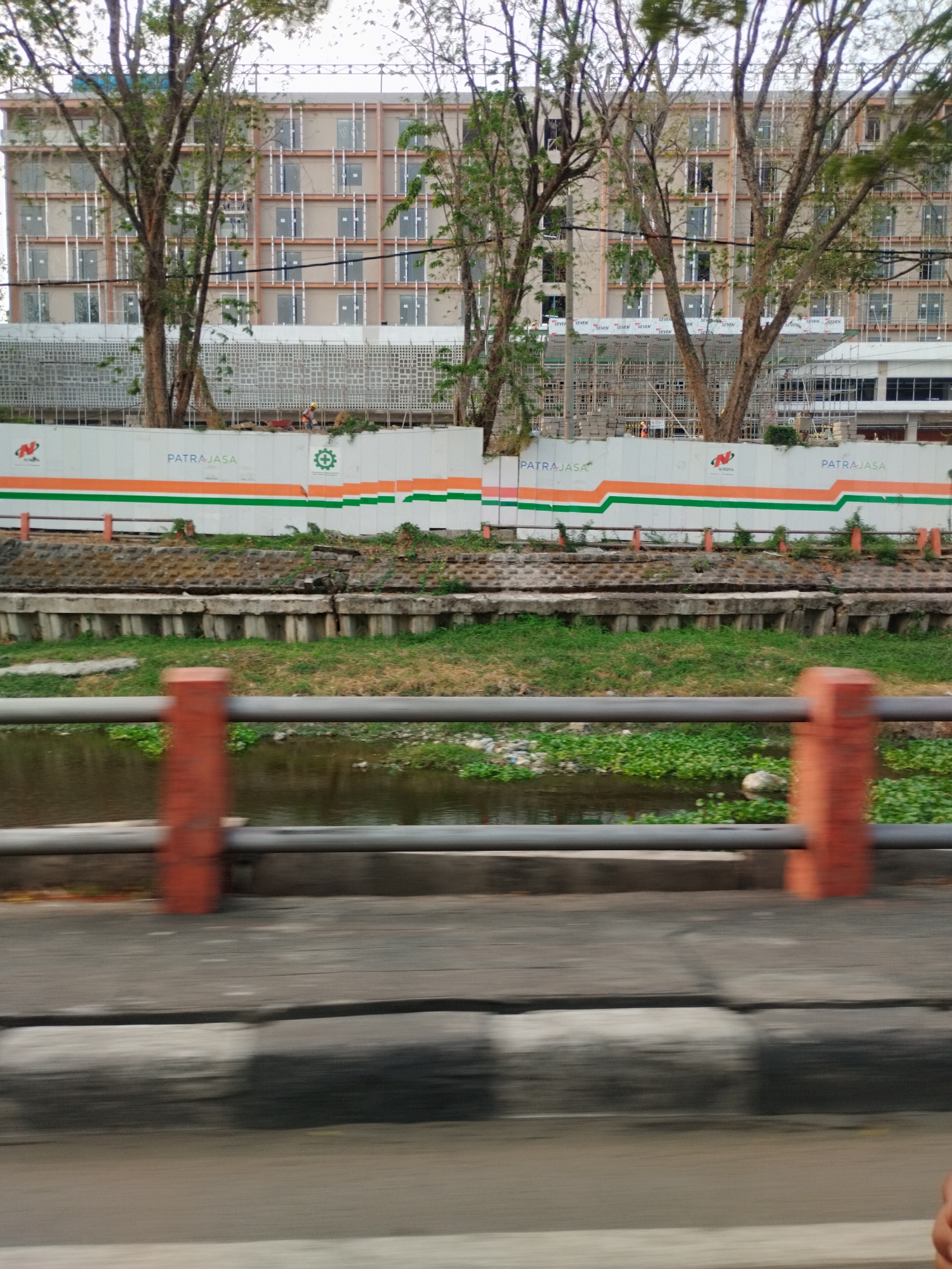 This is a medium-angle, outdoor, eye-level shot of a construction site adjacent to a river in Surabaya, Indonesia. In the background, a multi-story building is under construction, with scaffolding covering its facade and its windows mostly covered with white coverings. A white construction fence with orange and green stripes runs across the middle ground, with the words "PATRAJASA" printed multiple times on the fence. There is a single construction worker visible on the fence, pushing a wheelbarrow. In the foreground, a river flows with green aquatic plants covering its surface. The riverbank is reinforced with concrete structures and lined with grass. A metal railing with reddish-orange support posts runs along the riverbank. The bottom of the image shows a blurred view of a road or walkway with visible cracks.