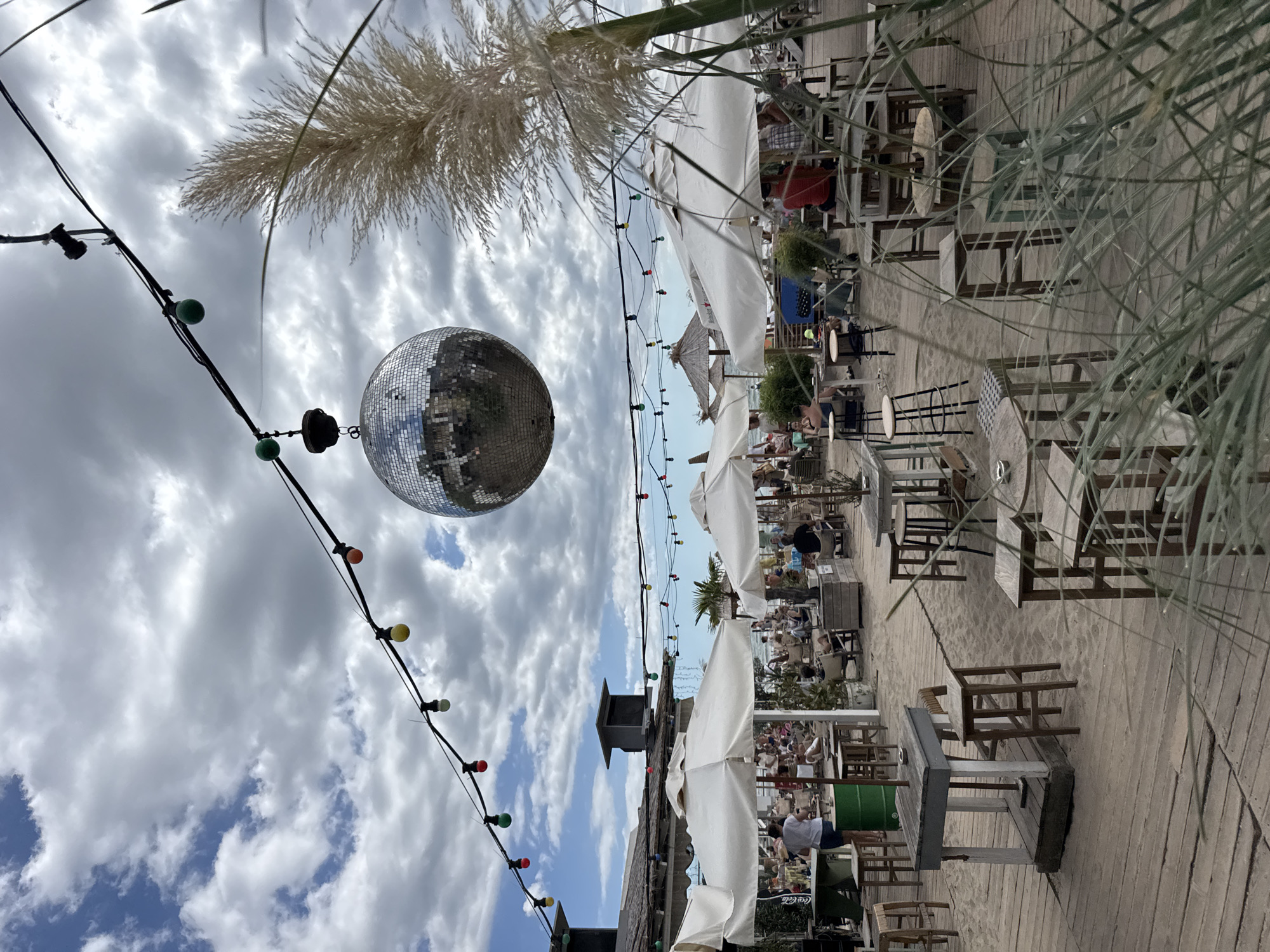 The image depicts a beachside bar or restaurant on a cloudy day. A large disco ball hangs from a string of colorful lights strung across the scene. In the foreground, there are wooden tables and stools set up on a sandy ground with wooden decking. People are seated at tables and lounging under white umbrellas in the background, suggesting a lively atmosphere. The location is identified as a beach in Varna, Bulgaria, likely a popular tourist spot. The overall impression is of a casual, festive outdoor venue preparing for or enjoying a day of leisure.