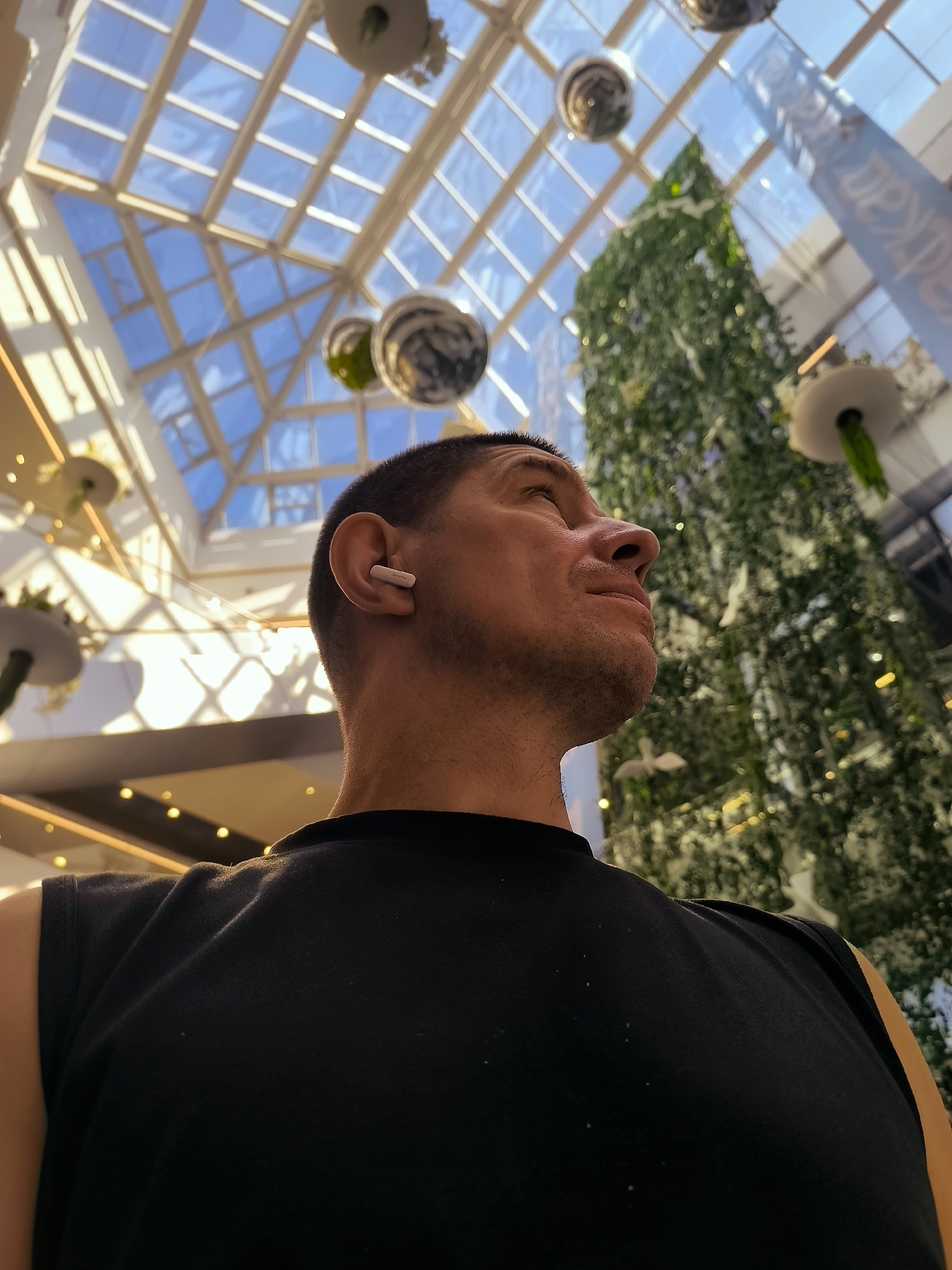 A man with short hair and a black sleeveless shirt looks upwards to the right of the frame. He is wearing a white wireless earbud in his right ear. In the background, a large, green, artificial Christmas tree is decorated with white ornaments and small yellow lights. Above the tree is a glass ceiling with geometric patterns, allowing sunlight to filter through. Several decorative spherical objects are suspended from the ceiling, and there are also some hanging plants. The scene appears to be indoors, possibly in a shopping mall or public space, in Qarağandı city administration, Kazakhstan.