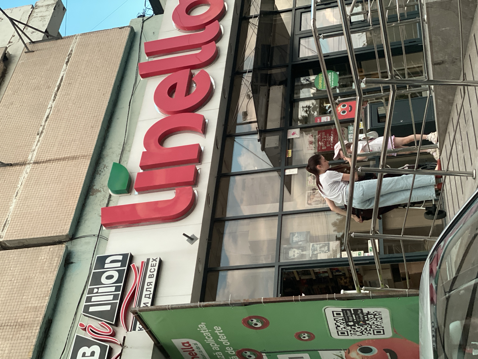The image shows the exterior of a "Linella" supermarket in Chișinău, Moldova. A large red "Linella" sign is prominently displayed above the entrance. To the left of the main sign, a smaller black and white sign reads "Billion" with a stylized "i" within the word. Below the "Billion" sign, another sign in Russian states "КОМФОРТ И ДЛЯ ВСЕХ" (Comfort for everyone). A green banner on the left side of the building advertises "Linella" with a QR code and mentions downloading the application.
Two individuals are visible near the entrance: an adult woman and a young child. The woman, wearing a white t-shirt and light blue jeans, is facing the child. The child, wearing a white t-shirt and a pink skirt or shorts, is looking upwards towards the woman. They are standing on a ramp with metal railings. Behind them, the entrance to the supermarket is visible through large glass windows. Several posters and advertisements are displayed on the windows. A red cartoon-like apple character with eyes and a mouth is also visible on the green banner. The building's facade features a tiled upper section. A silver car is partially visible in the foreground. The sky is blue and clear.