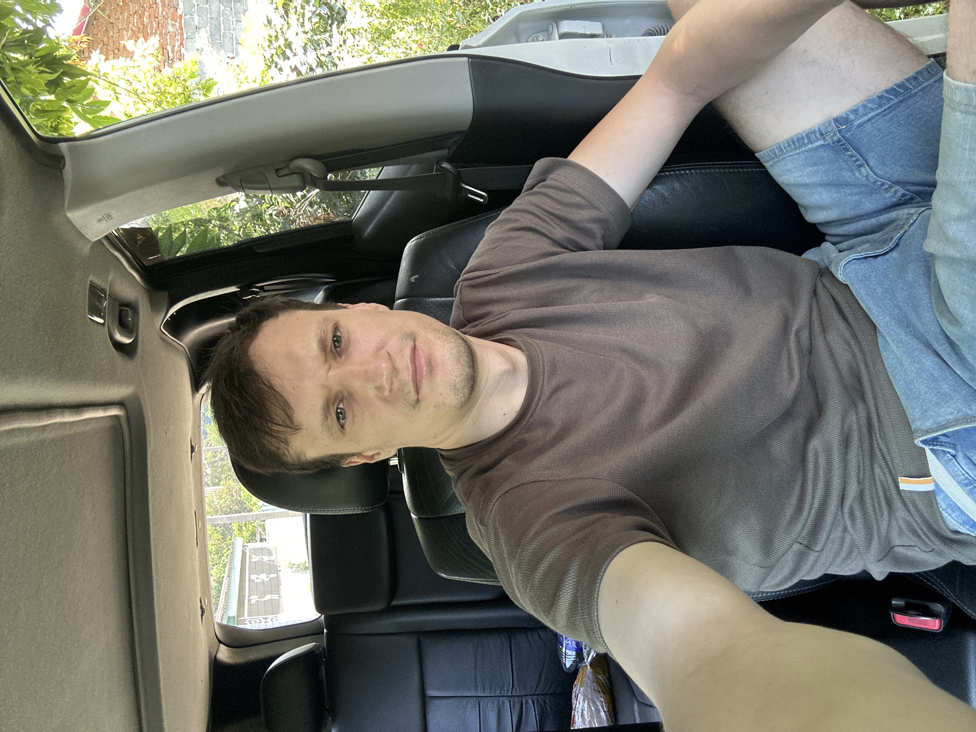 A man sits in the driver's seat of a car, looking at the camera. The interior of the car appears to be a dark leather. Through the open car door and the rear windows, we see a green, leafy environment with trees and vegetation. A brick wall is visible in the background. The man is wearing a brown t-shirt and denim shorts. The location is identified as Makiv, Ukraine.