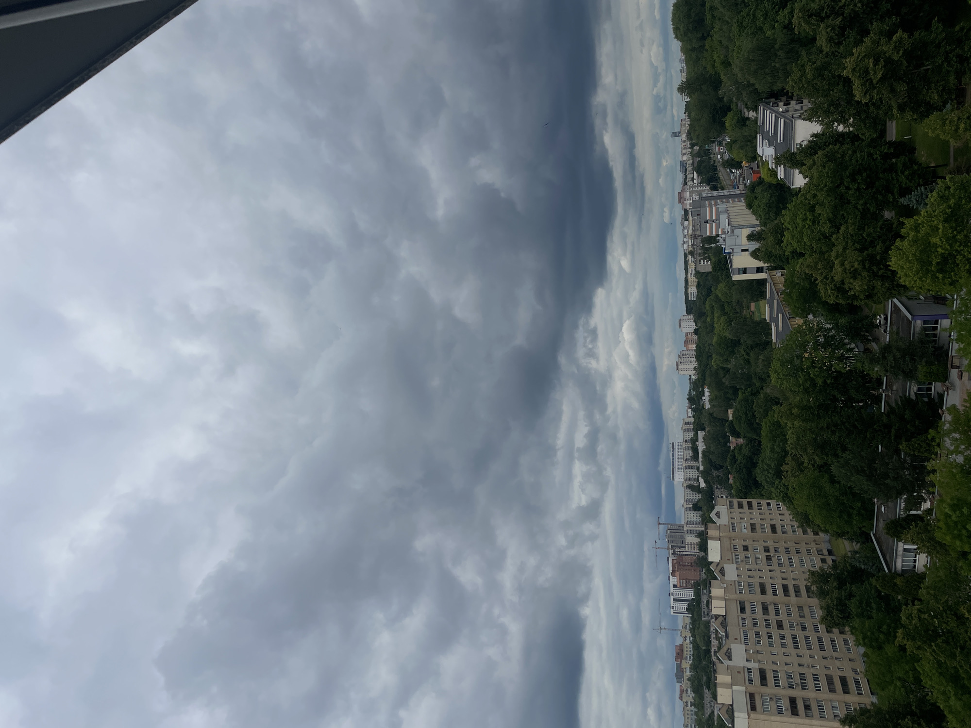 The image shows a panoramic view of a city with a cloudy sky overhead. In the foreground, there is a large apartment building. Behind it, a dense forest with lush green trees stretches across the landscape. Further in the distance, more buildings and construction cranes are visible, suggesting ongoing development in the city. The overall impression is that of a city that is a blend of urban and natural environments. The scene appears to be from a high vantage point, possibly a balcony or a tall building, looking out over the cityscape. The sky is filled with dark, heavy clouds, indicating a possibility of rain or a stormy weather. The presence of trees and buildings suggests it is a populated area within a city. The architecture of the buildings appears to be of a modern style, common in many European cities. The city is identified as Minsk, Belarus.