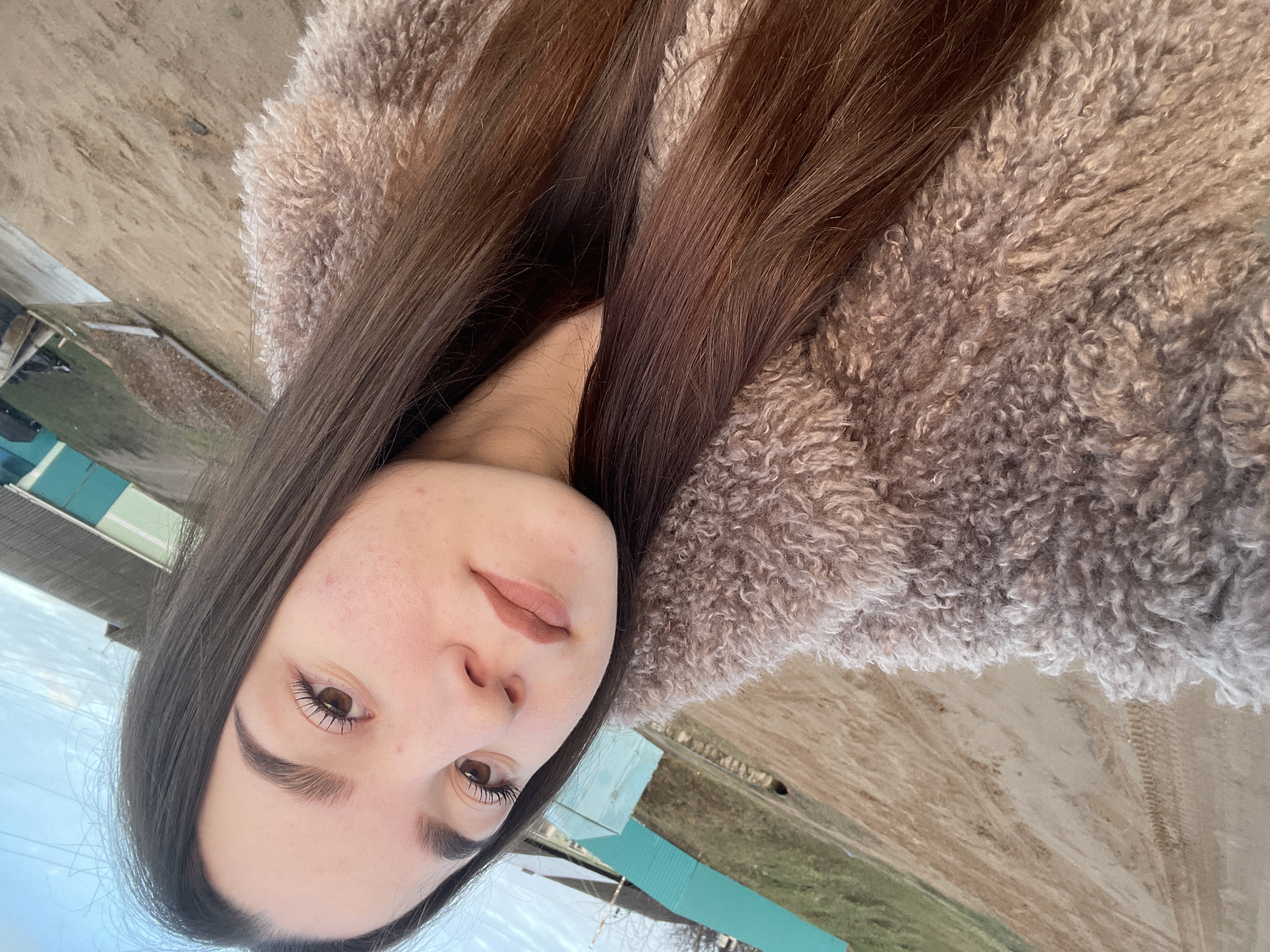 A young woman with long brown hair, wearing a furry light brown jacket, is the main subject of a selfie. Her face is tilted towards the camera, showing brown eyes, defined eyebrows, light skin, and pink lips. The background shows a rural setting with a visible building, possibly a house, with light blue and green elements. There is a sandy or earthy ground visible, suggesting an outdoor location, possibly in Ольховка, Russia, but the specific details that would confirm this location are missing. The image appears to be taken during the day.