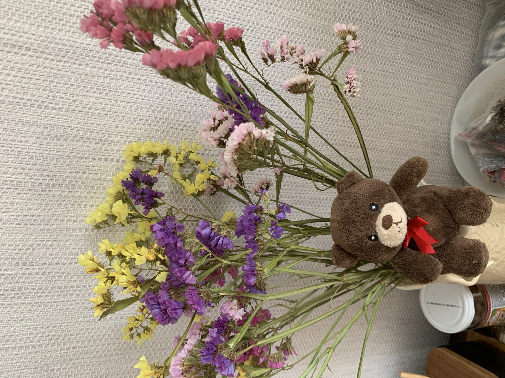 In Kapyĺ, Belarus, a small, brown teddy bear with a red ribbon is positioned in front of a vase of dried flowers. The flowers are a mix of yellow, purple, and pink hues, with some appearing lighter and more delicate than others. The background is a textured, light-colored wall. To the left of the teddy bear and vase, a white-lidded jar is partially visible. Below the flowers and to the right, a white plate holds various small items. The overall scene suggests a domestic setting, possibly a tabletop or shelf.